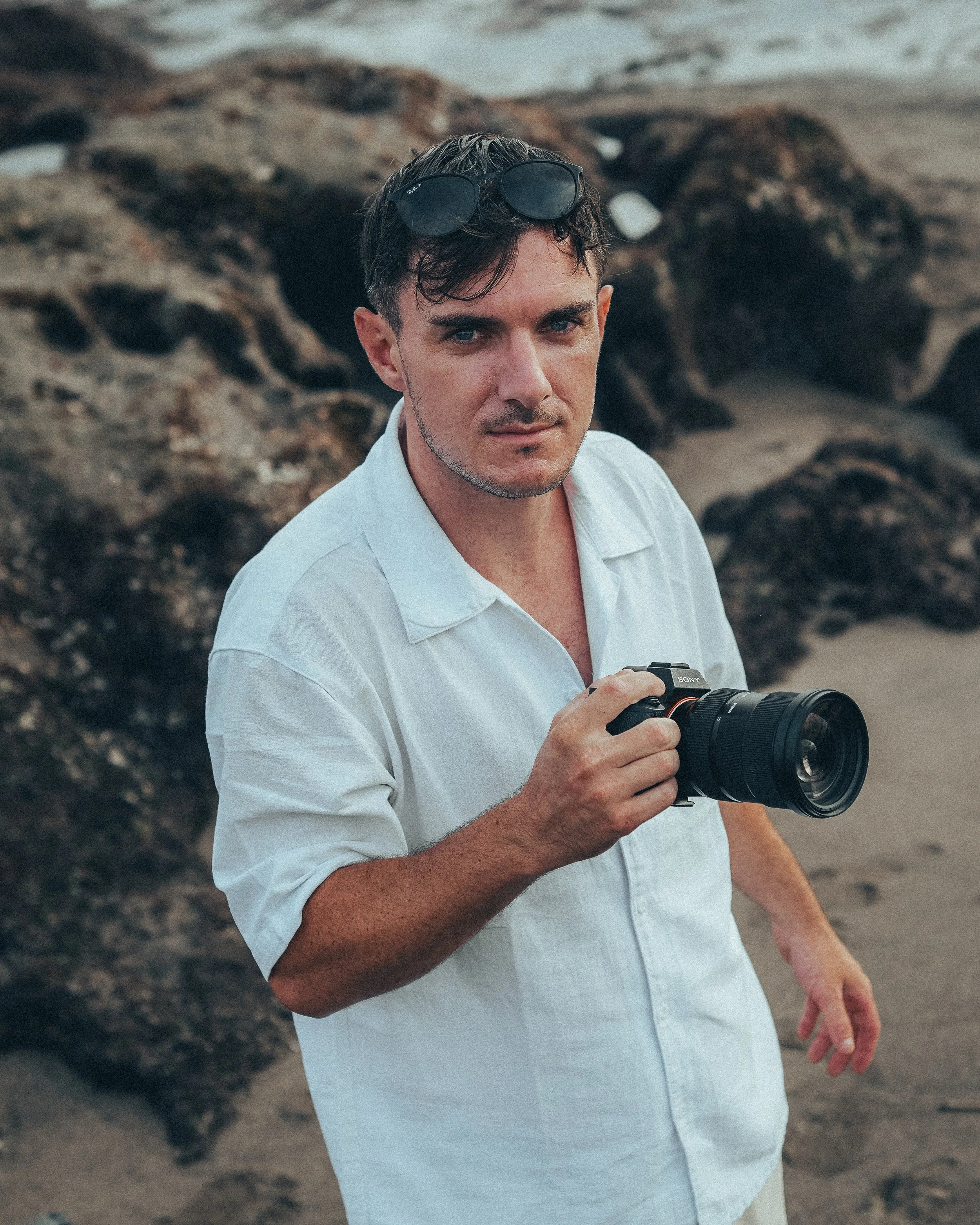 Un homme tenant un appareil photo sur une plage rocheuse, portant une chemise blanche et des lunettes de soleil sur la tête.