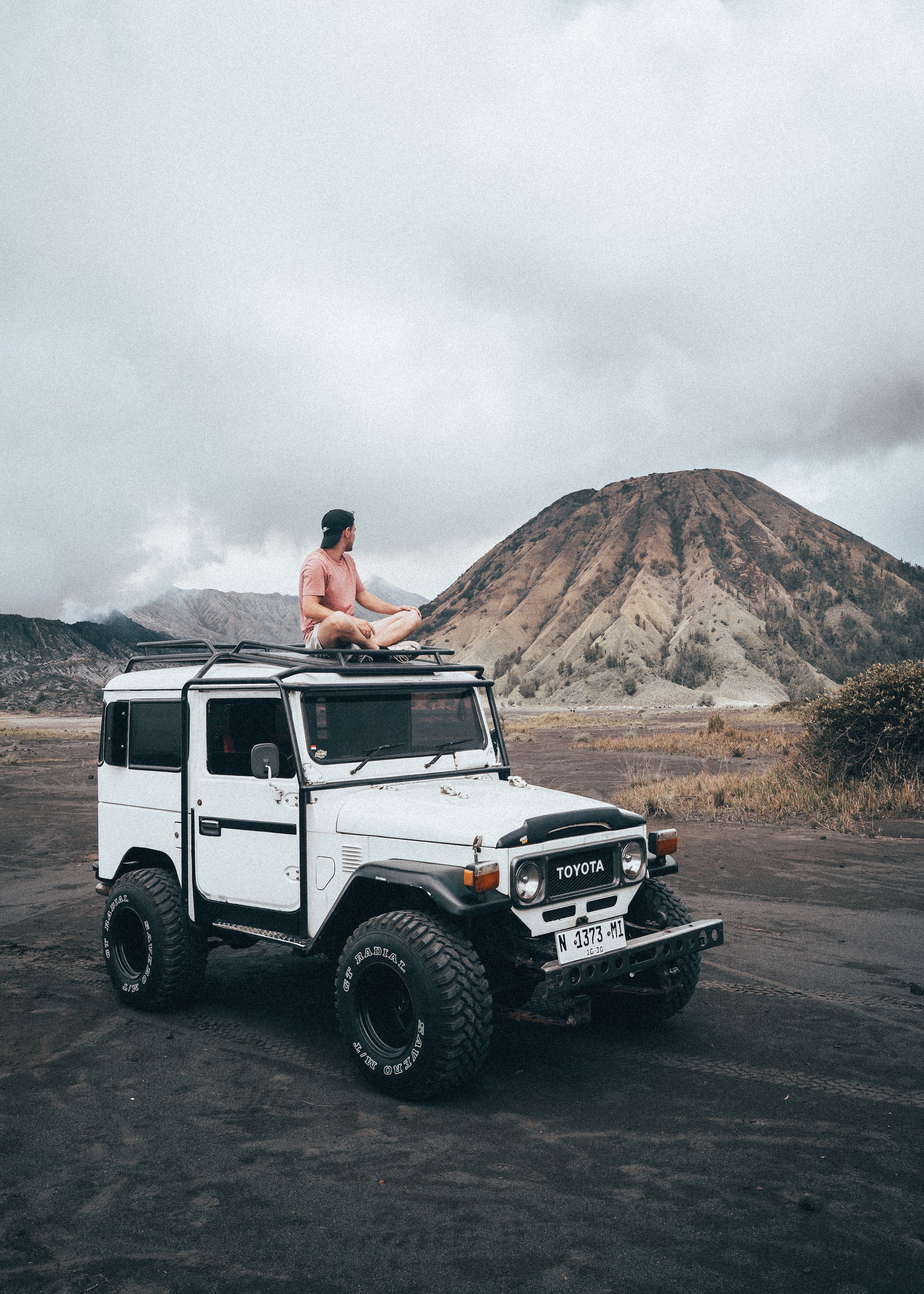 Un homme assis méditant sur le toit d'un véhicule tout-terrain blanc, avec un volcan en arrière-plan dans un paysage aride, ciel nuageux.