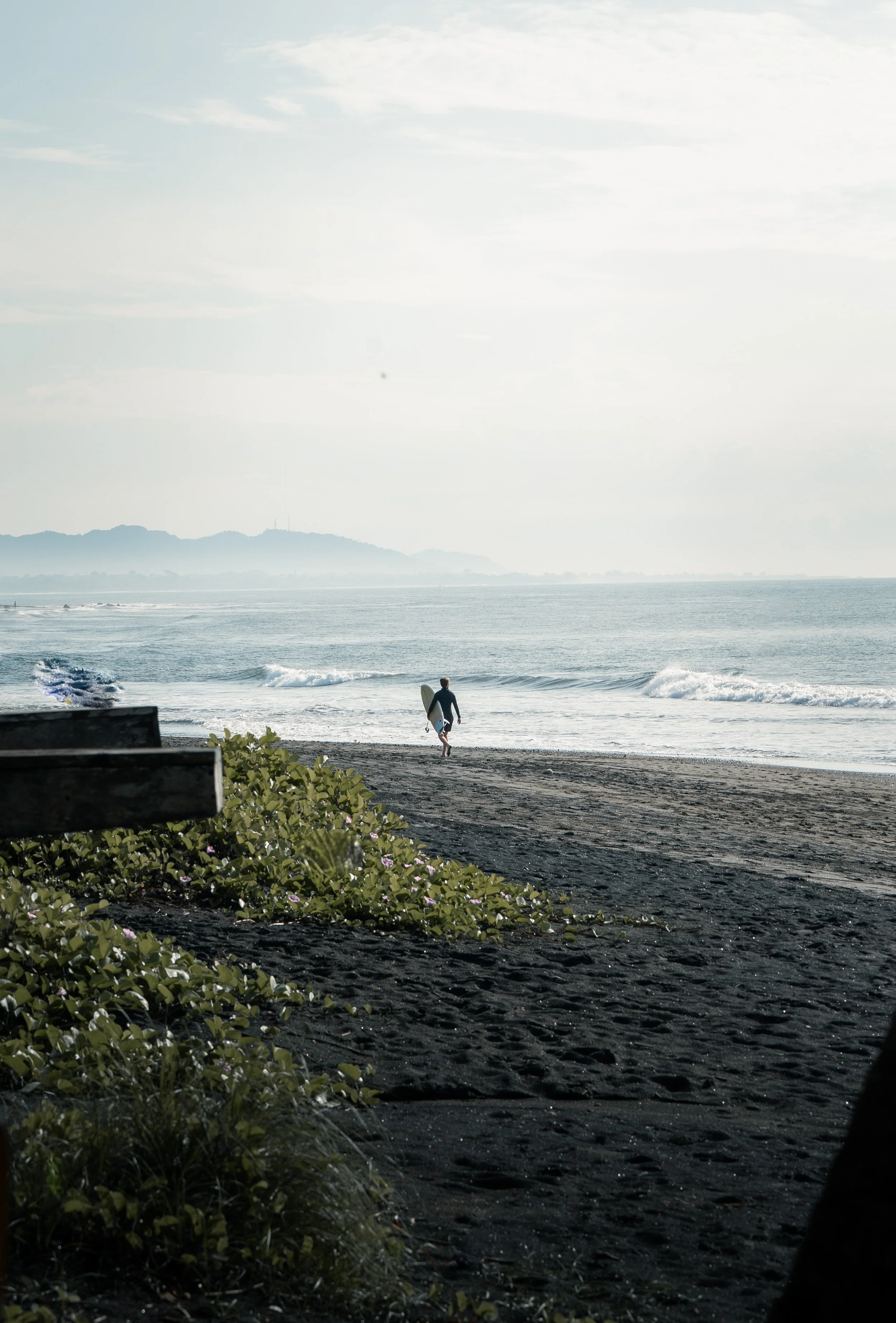 Un surfeur marche vers l'océan sur une plage de sable noir, avec des vagues et des montagnes en arrière-plan, sous un ciel partiellement nuageux.