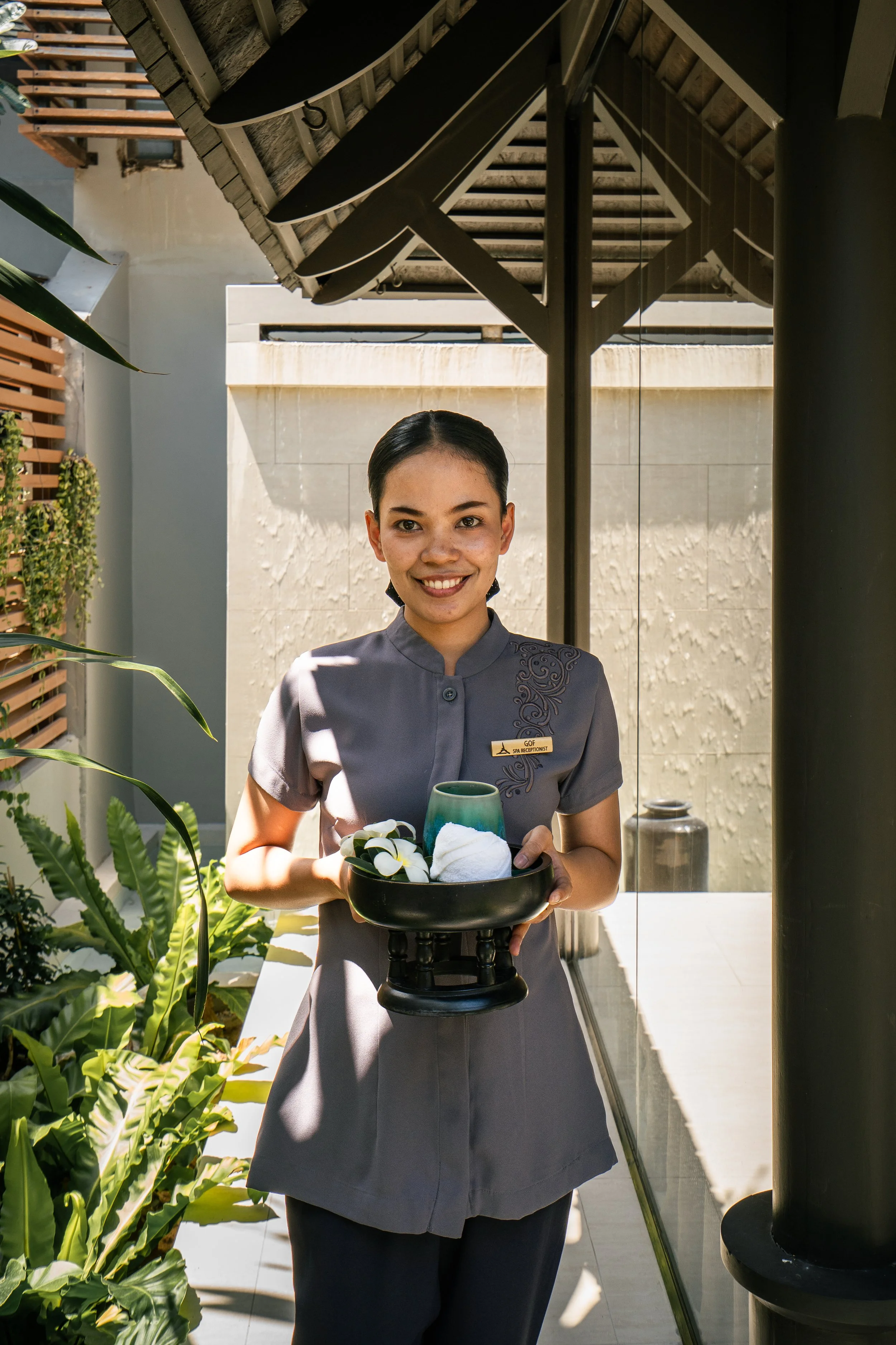Une femme souriante portant un uniforme gris foncé, tenant un plateau avec des serviettes et des fleurs, à l'extérieur d'un bâtiment moderne avec des plantes vertes.