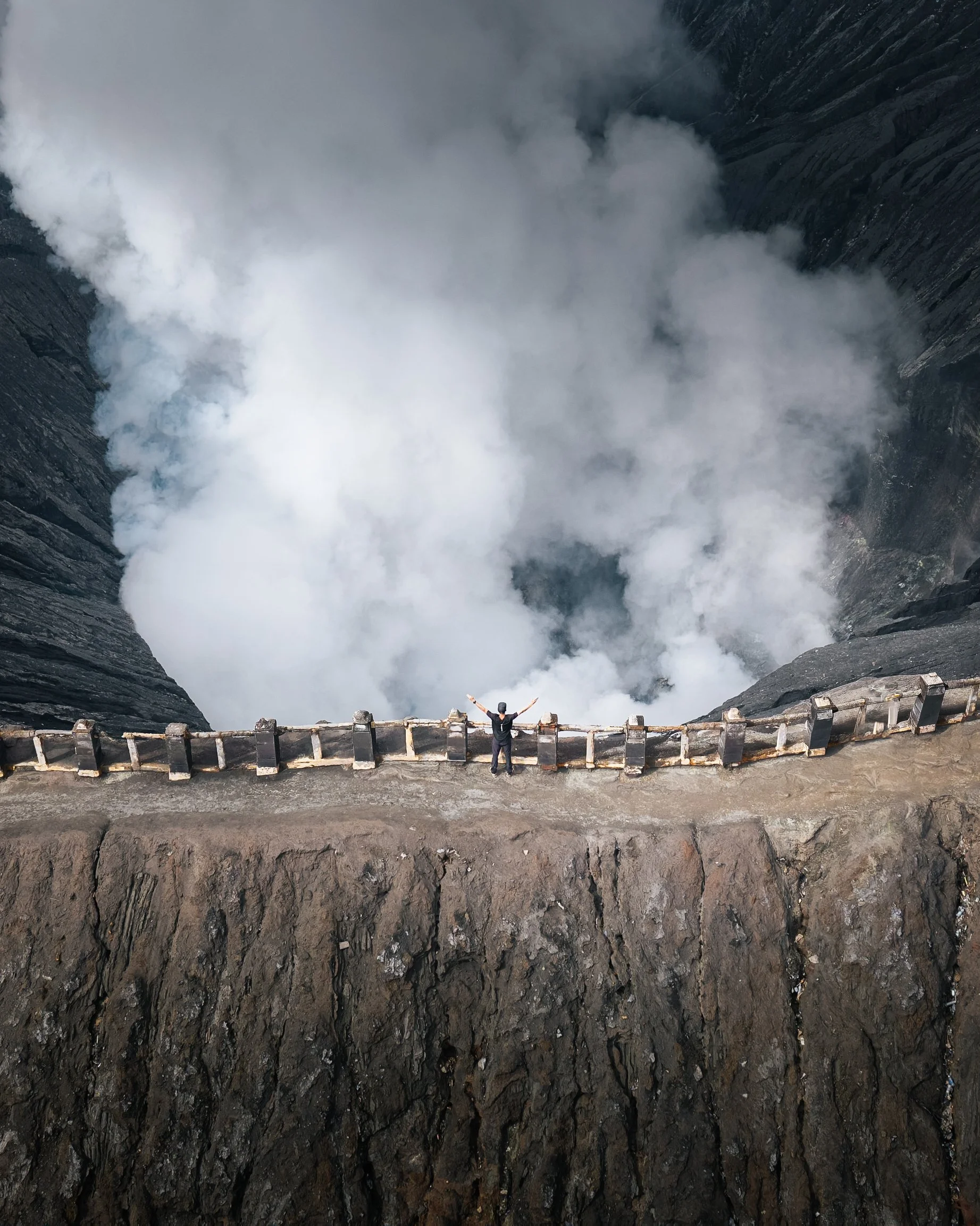 Une personne se tient devant un gouffre profond, avec des nuages de vapeur ou de fumée en haut, entourée de parois rocheuses sombres.