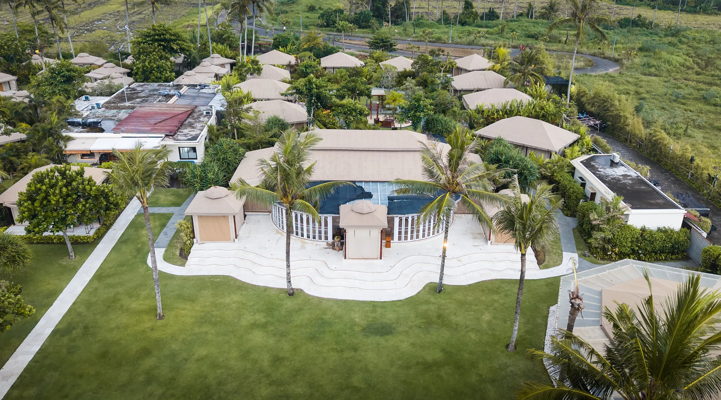Vue aérienne d'une maison de vacances entourée de palmiers, avec un jardin bien entretenu et plusieurs petites villas environnantes dans un environnement tropical.