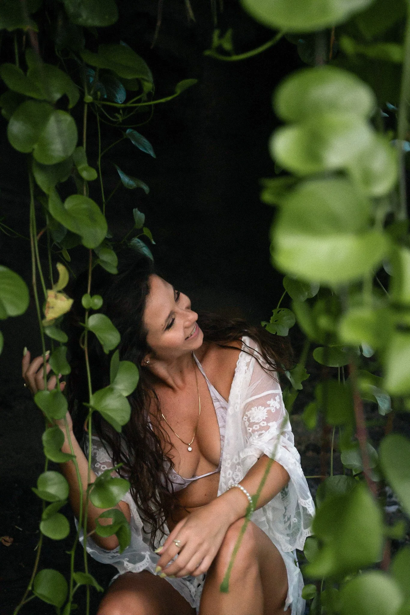 Une femme souriante avec des cheveux longs, portant une robe blanche en dentelle et un collier, assise parmi des feuilles vertes