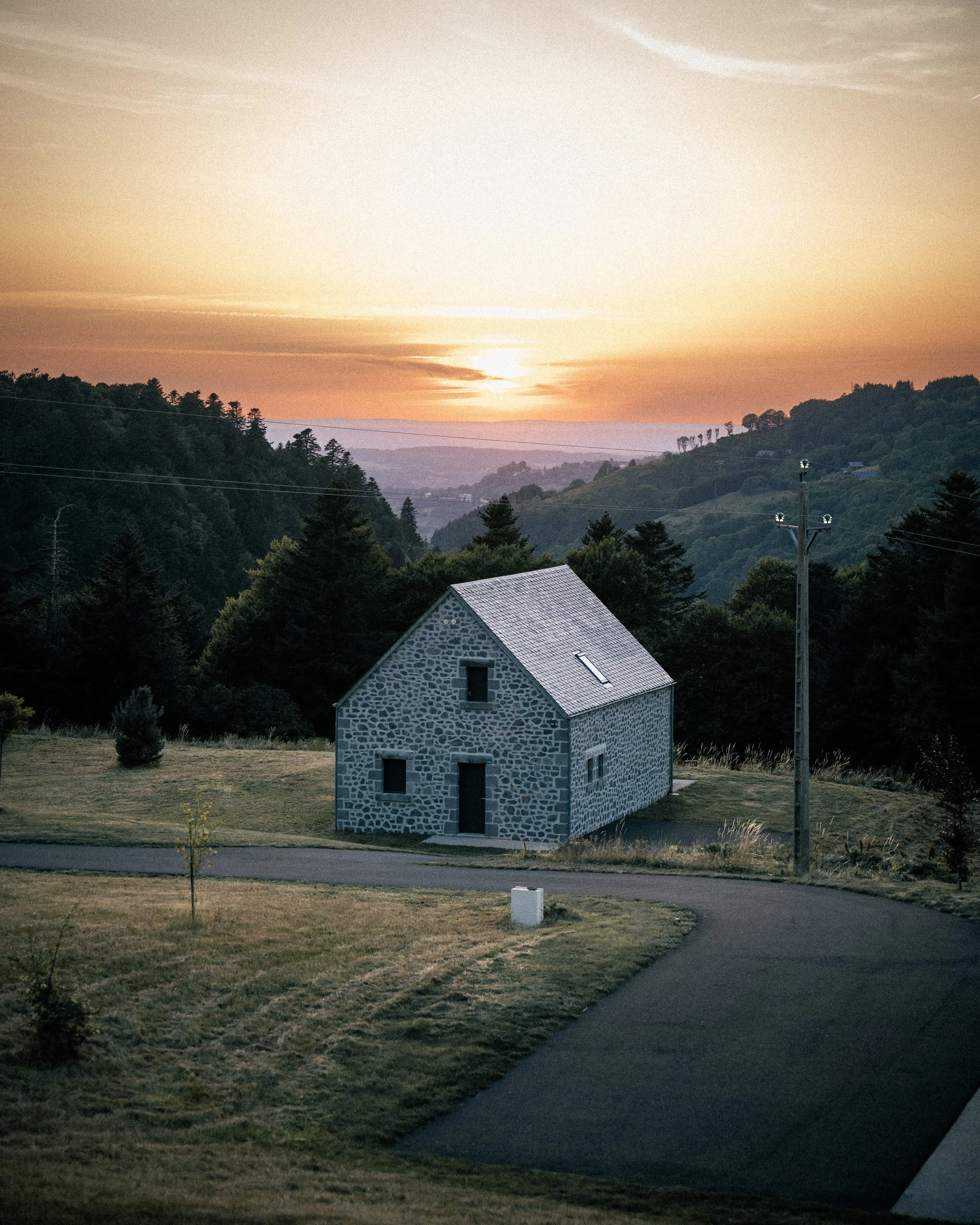 Une maison en pierre avec un toit en tuiles, située dans un paysage vallonné au coucher du soleil, entourée d'arbres et d'une route pavée.