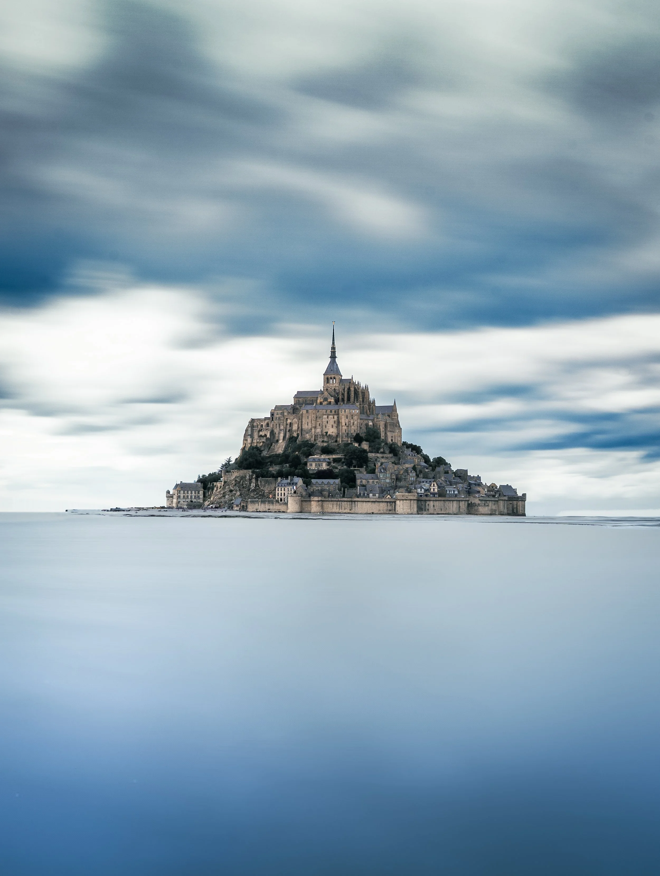Une photo du Mont Saint-Michel avec un ciel nuageux et une mer calme en premier plan.