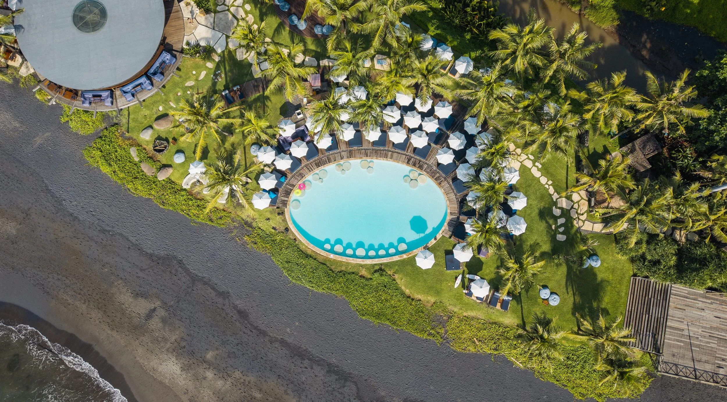 Vue aérienne d'une piscine en forme d'arc entourée de parasols et de chaises longues, avec des palmiers, un barbecue et un chemin de pierre, près d'une plage de sable et de l'océan.