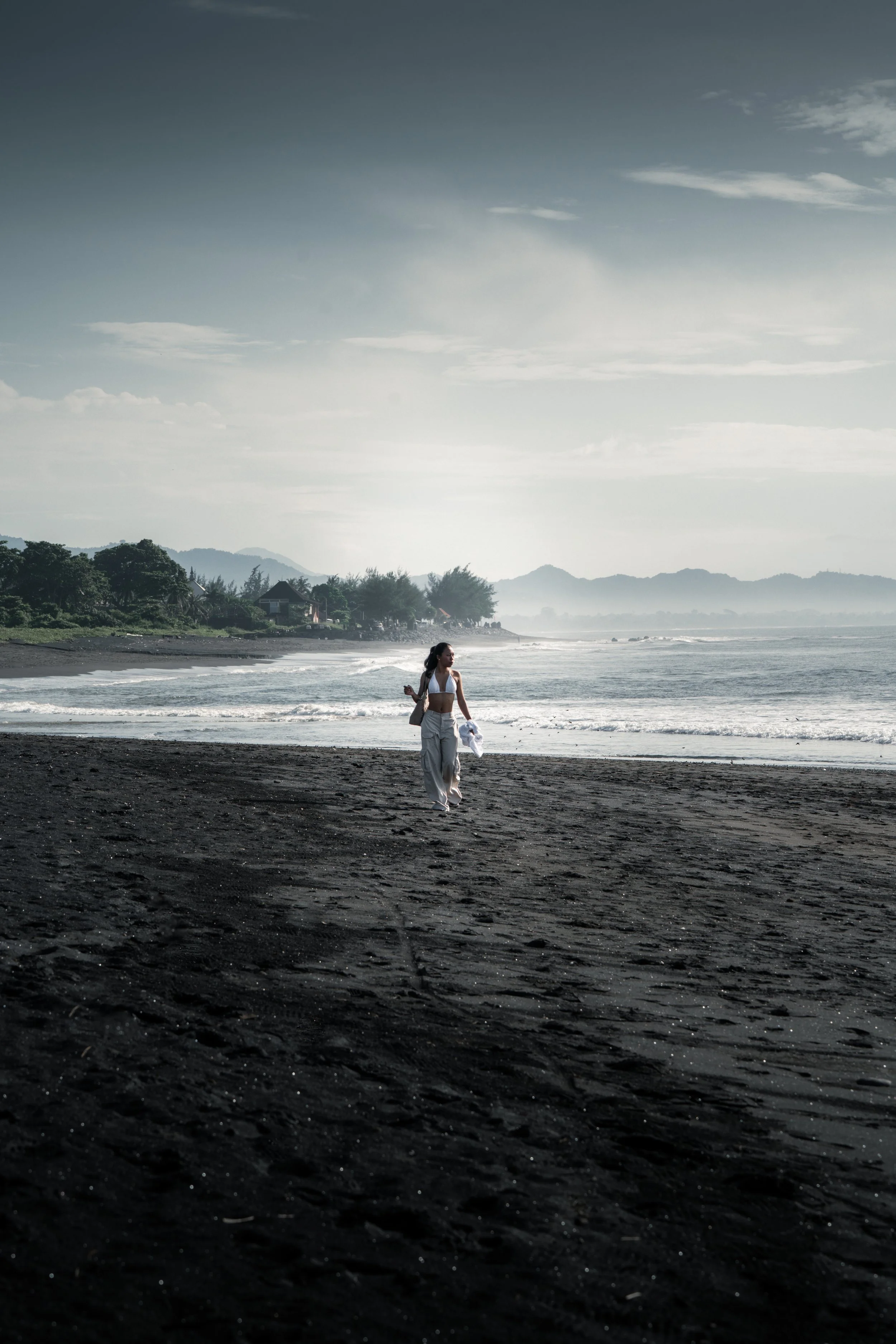 Femme marchant sur une plage de sable noire, regardant vers la mer avec des montagnes en arrière-plan et un ciel partiellement nuageux.