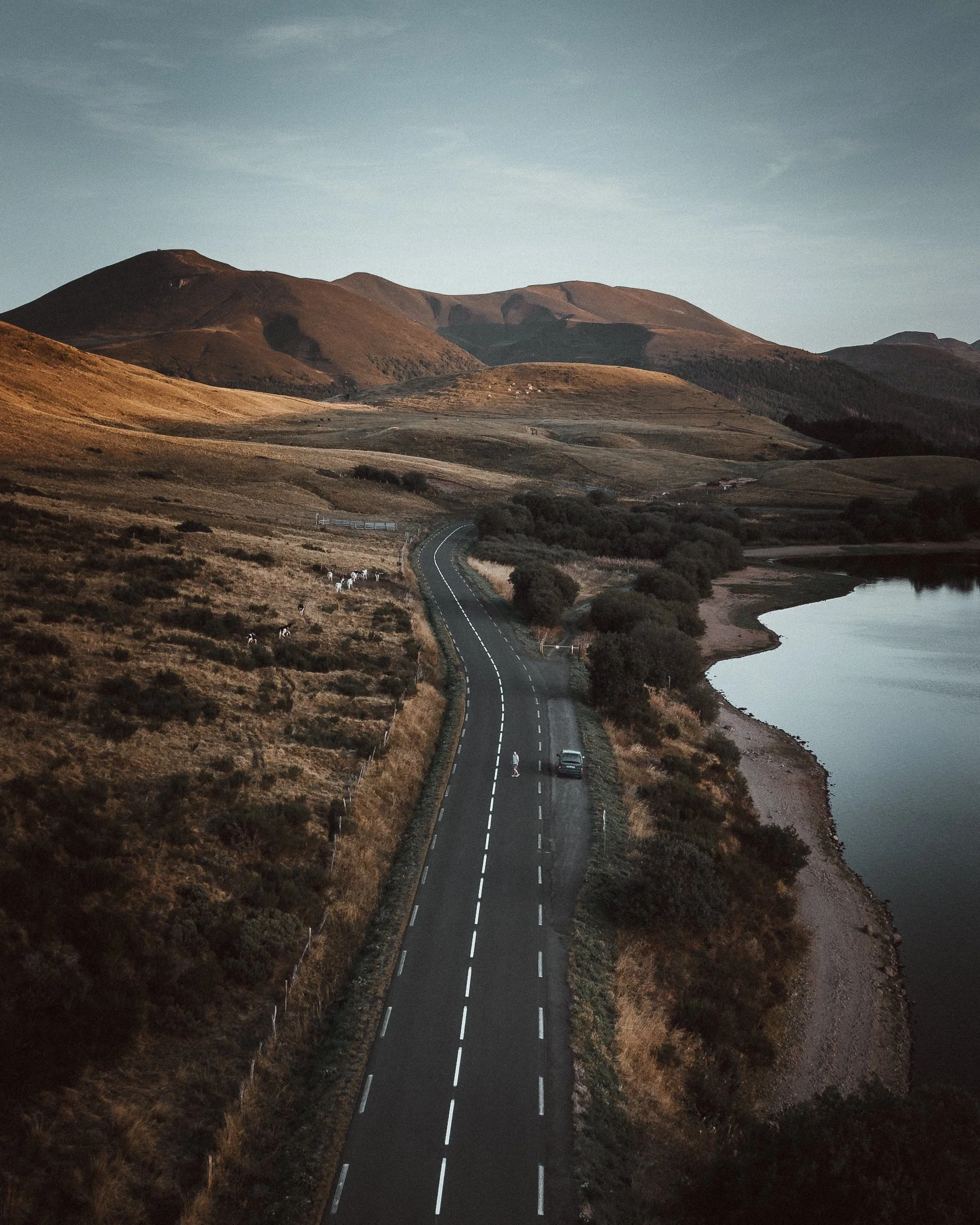Une route sinueuse longe un lac, entourée de collines et de montagnes, avec un ciel clair et peu de végétation.