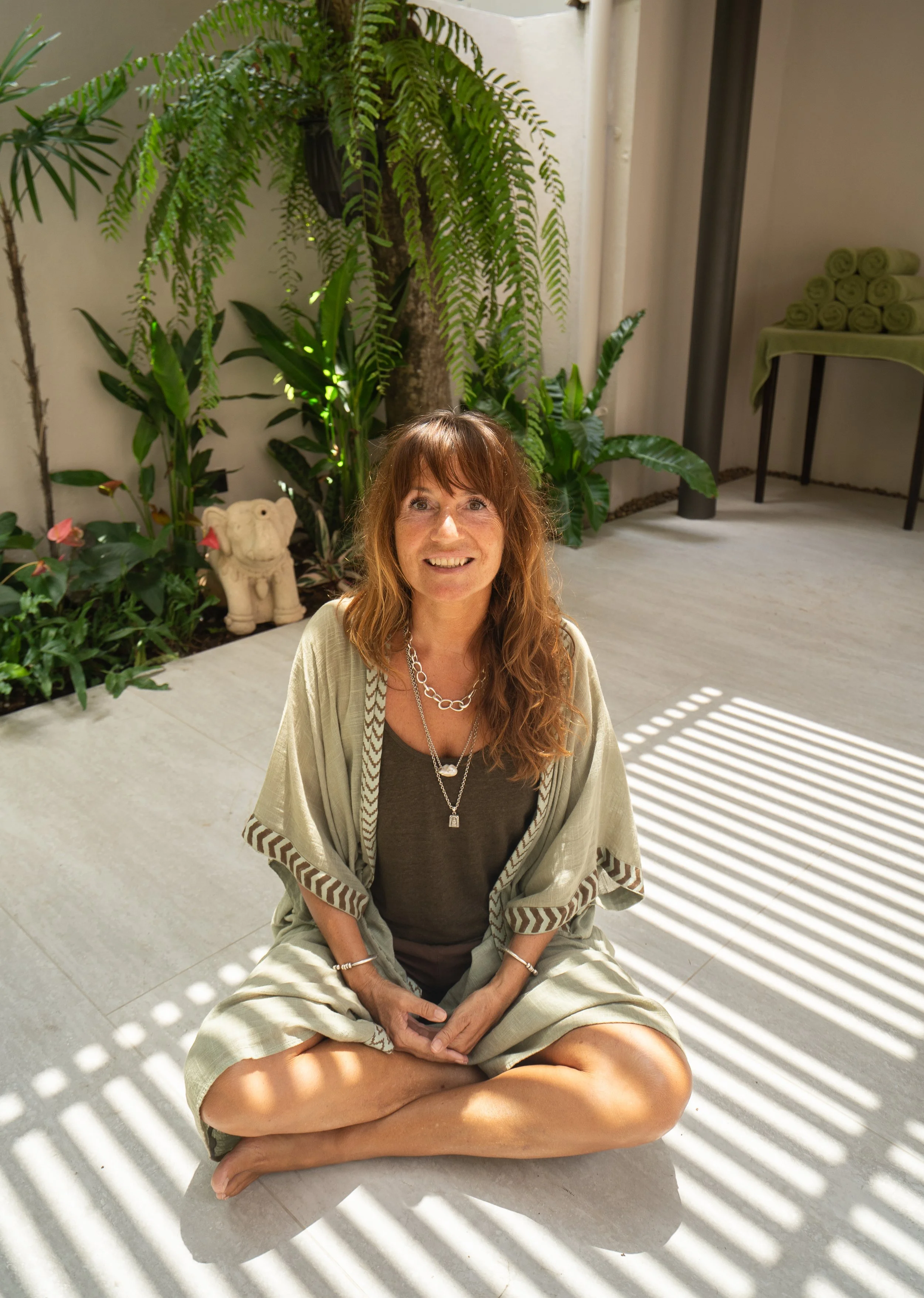 Femme assise au sol, souriante, entourée de plantes vertes, sous un éclairage de lumière naturelle filtrée par des ombres.
