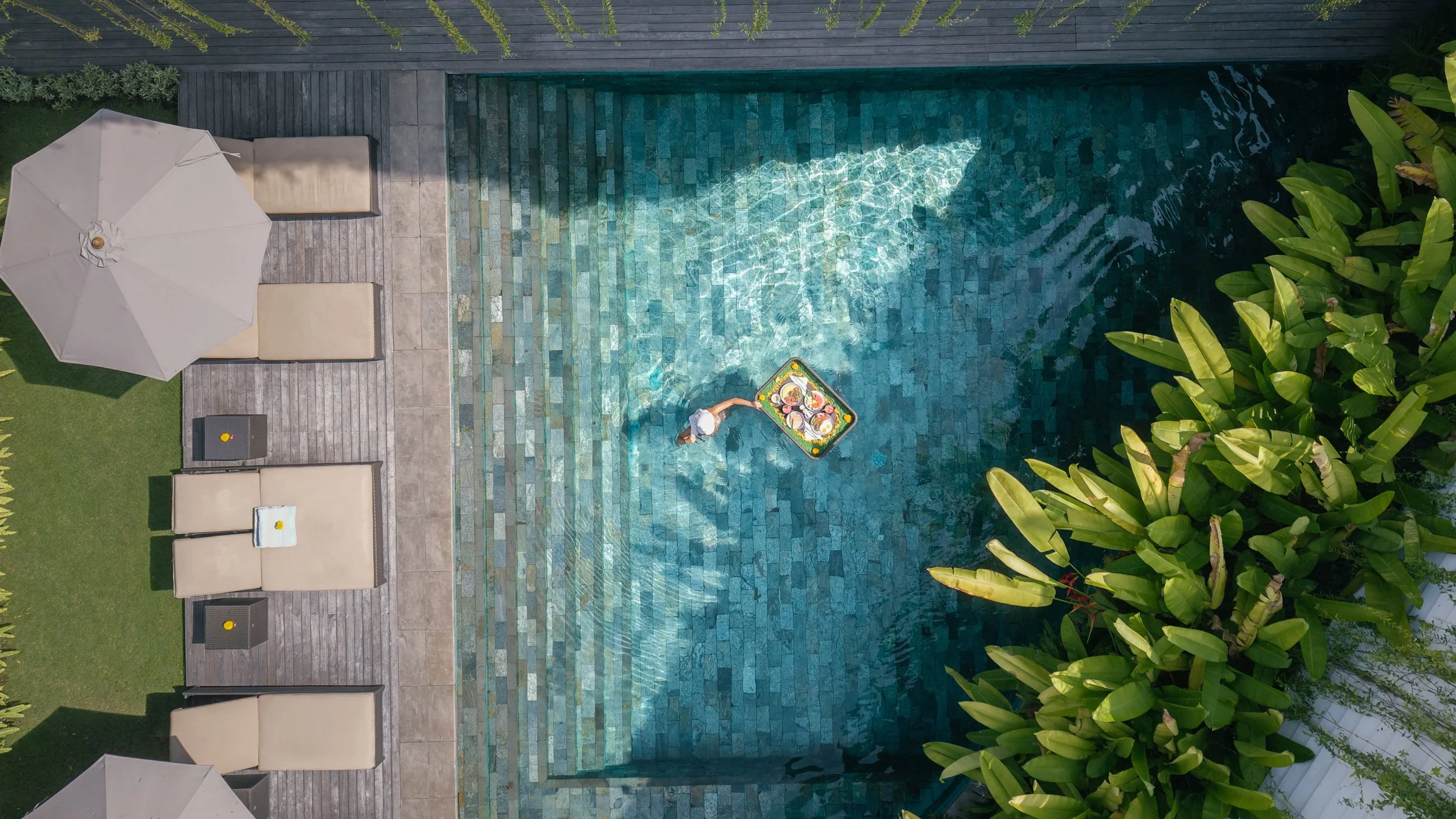 Vue aérienne d'une piscine en forme rectangulaire entourée de mobilier de plage, d'un grand parasol blanc, et d'une végétation luxuriante.