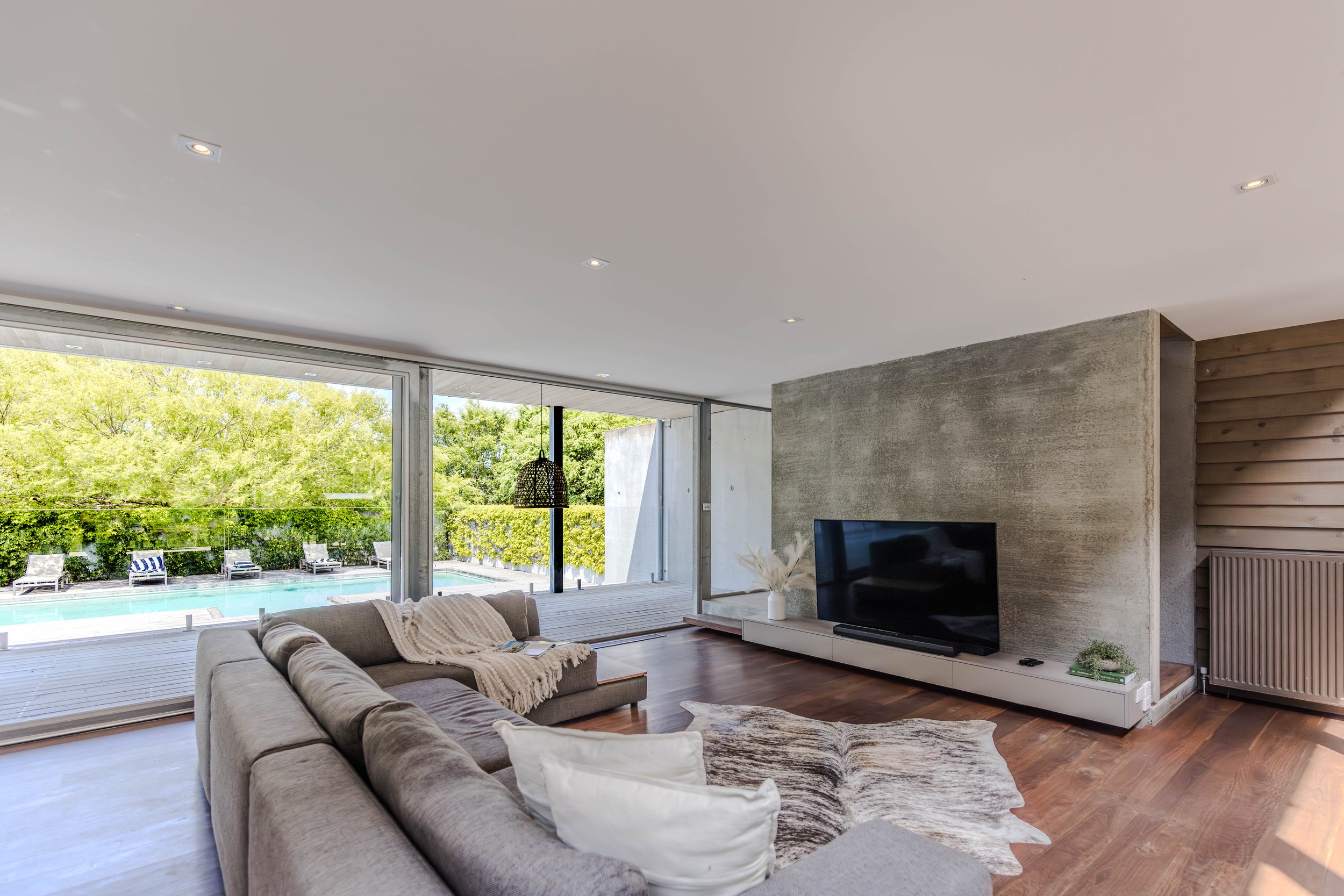 Modern living room with large sliding glass doors that open to a backyard with pool and lounge chairs. The room has a gray sectional sofa with white pillows and a cream throw blanket, a wooden floor, a cowhide rug, a TV on a white console, and a textured concrete accent wall.