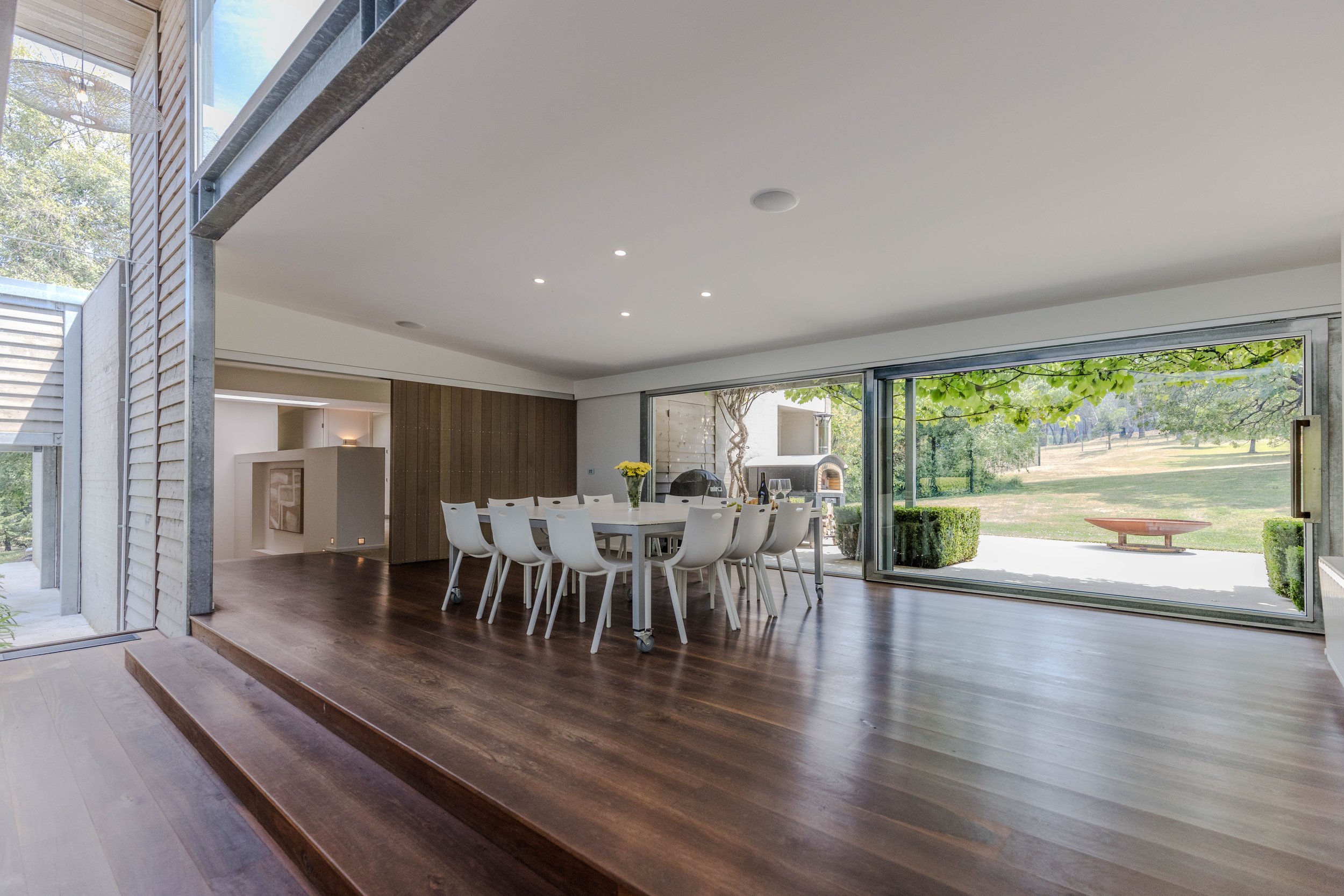 Modern dining room with large sliding glass doors overlooking a green outdoor space, white dining table with 12 chairs, wooden flooring, recessed ceiling lights, and a bouquet of yellow flowers on the table.