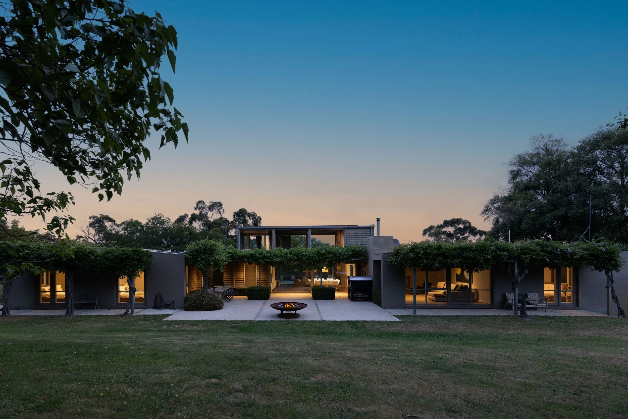 Modern house in Mount Macedon with large windows and a central courtyard, surrounded by trees, at dusk with the sky transitioning from light to dark.
