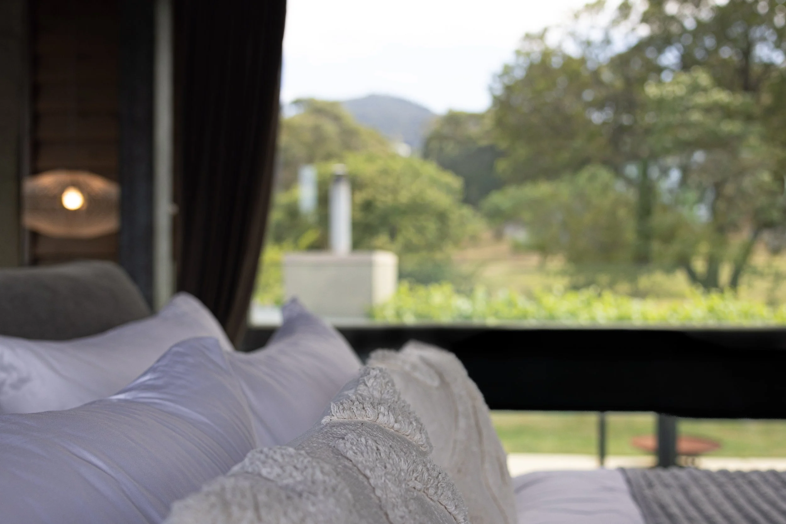 A cozy bed with white pillows and a textured cream blanket, overlooking a blurred outdoor landscape with green trees and hills through a large window.
