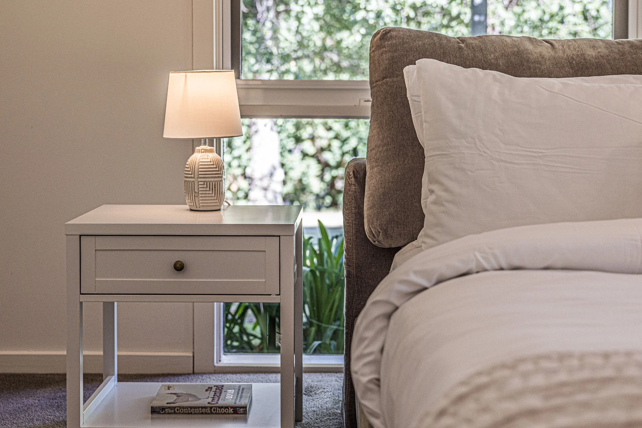 A bedroom with a white bedside table and a beige upholstered bed with white bedding. There is a lamp with a patterned base on the table, and a window showing green foliage outside.