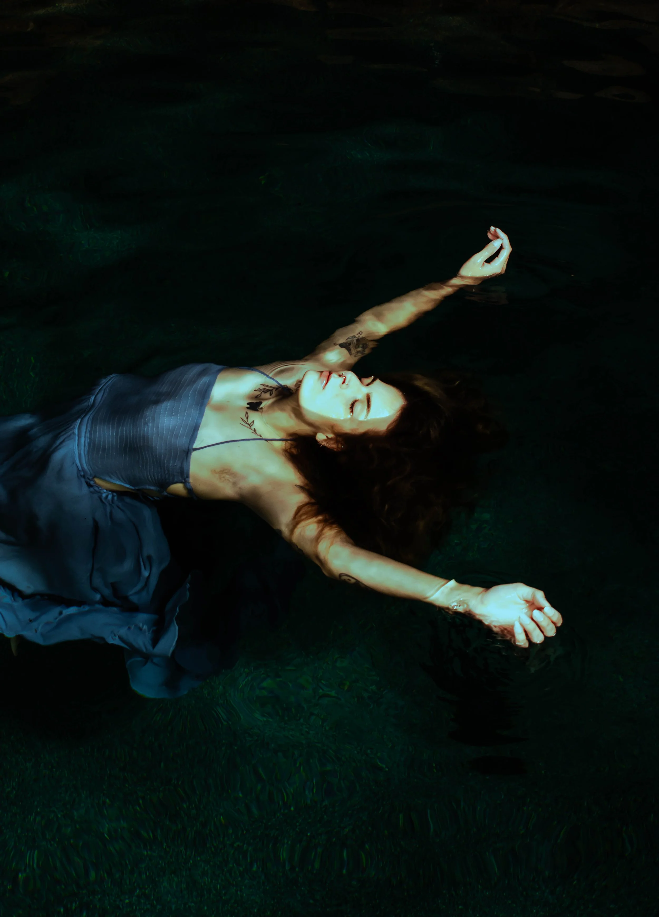 A woman with tattoos and long dark hair is floating in dark water with her eyes closed, wearing a blue dress, with arms stretched out and a peaceful expression.