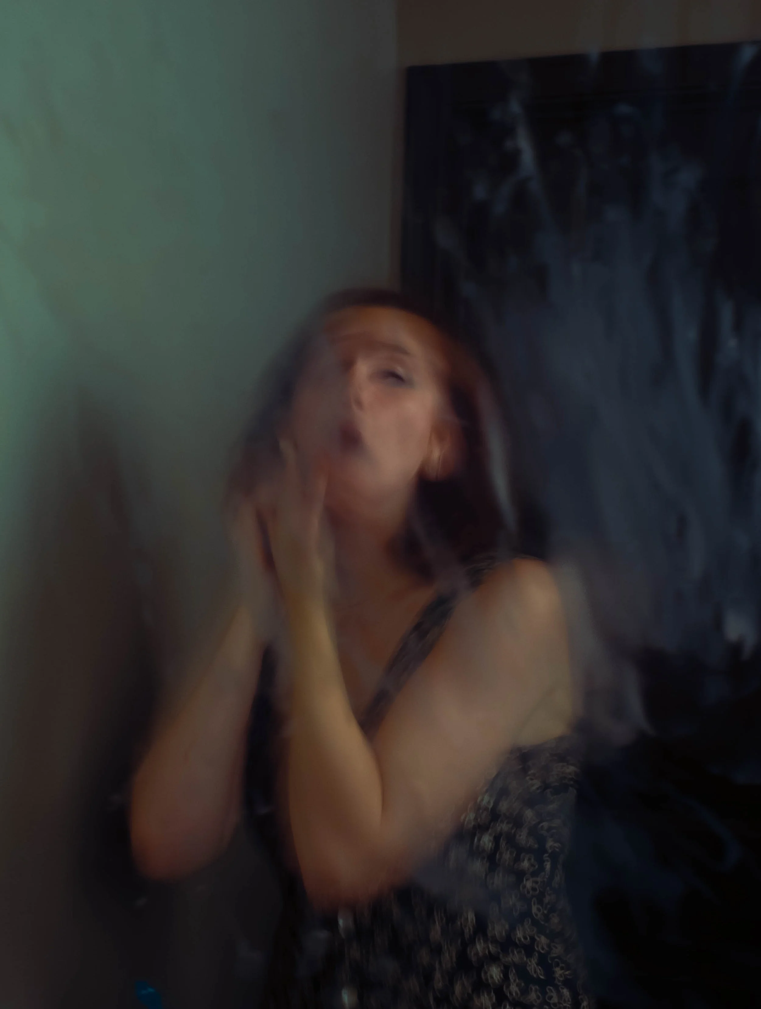 A woman with red hair in a blurry, motioned shot, posing with her hands near her face in a dimly lit room.