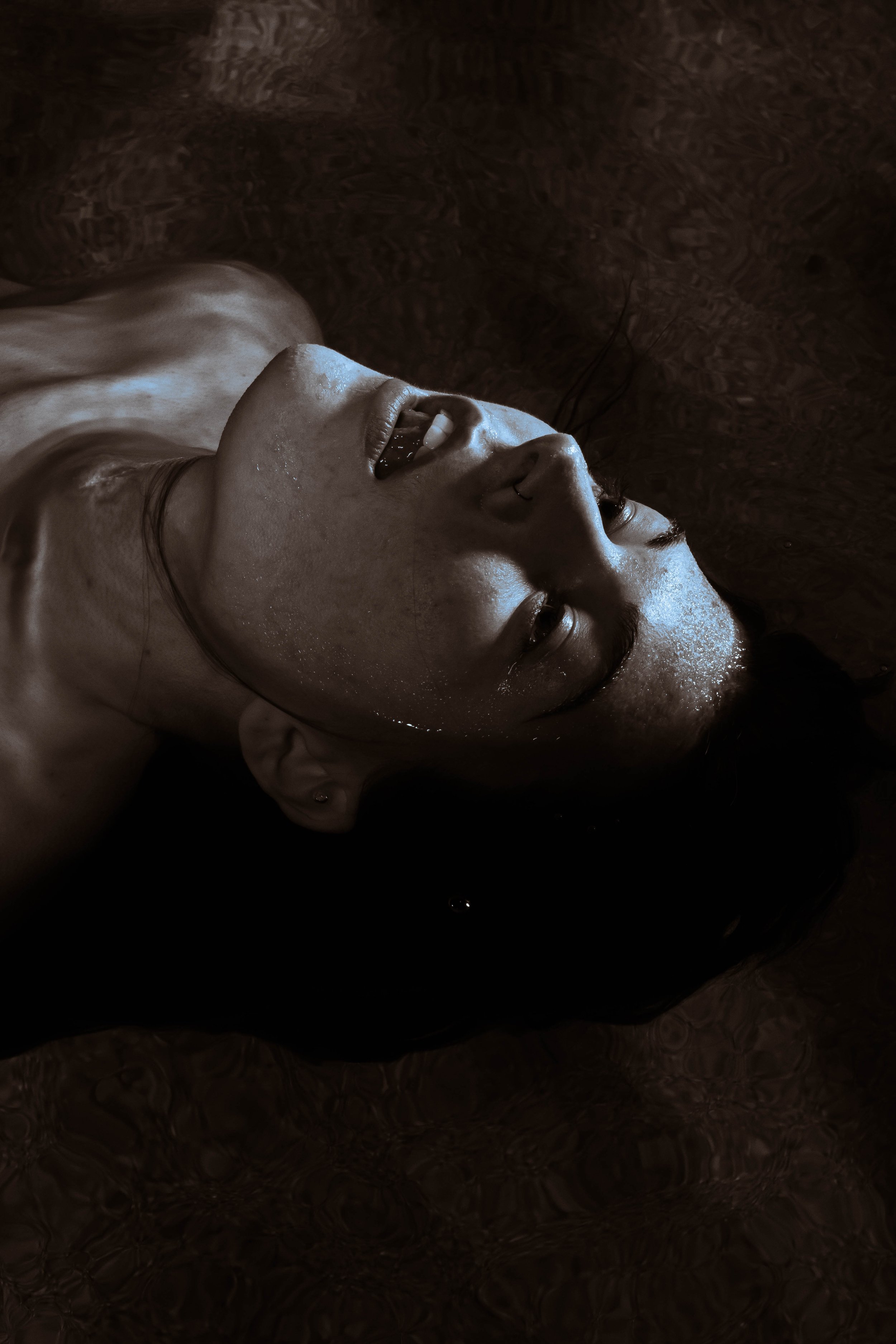 A woman with wet hair and face, lying in water, looking up with a serene expression.