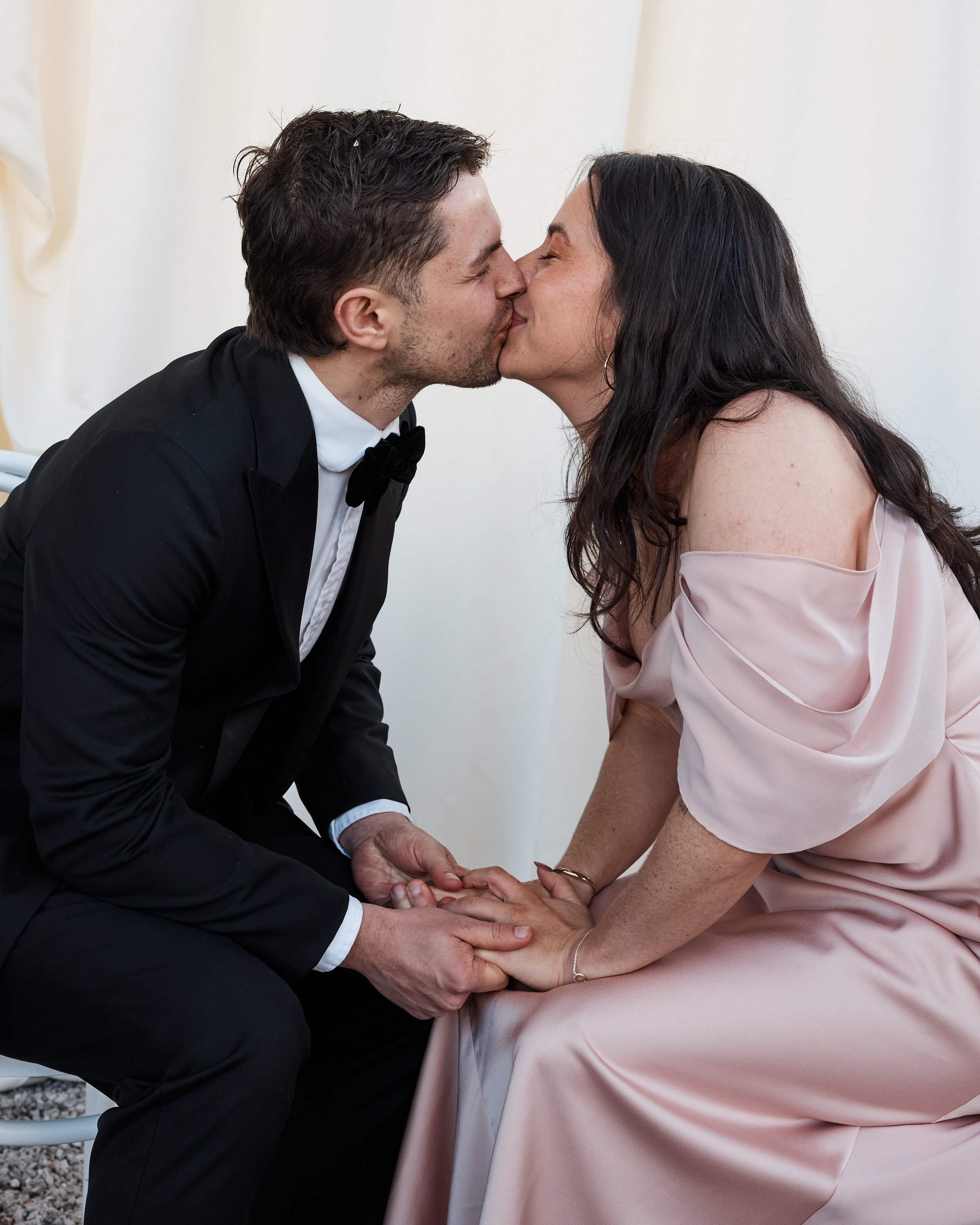 A man in a black tuxedo and a woman in a pink dress share a kiss while holding hands.