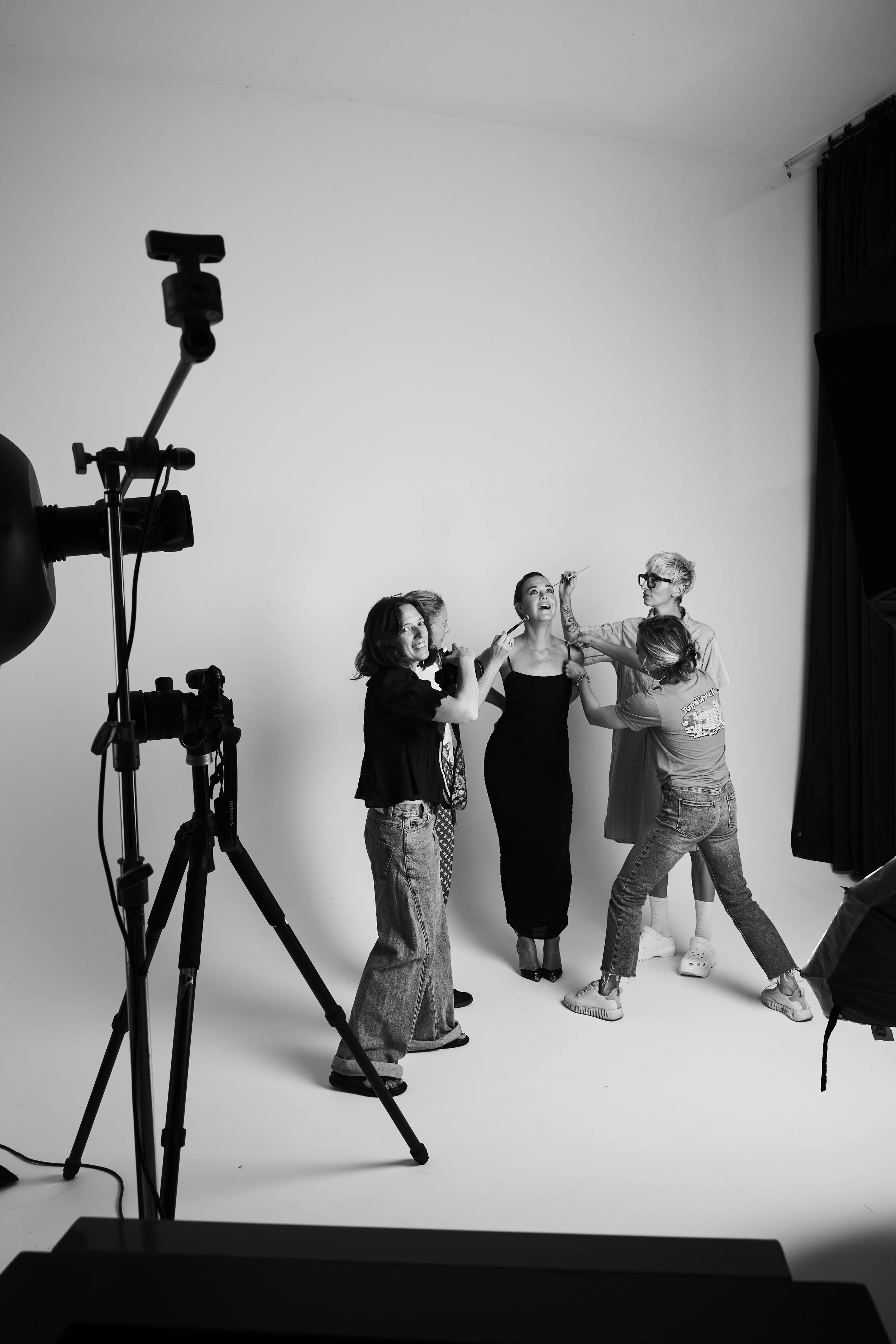 A black-and-white photo of women in a photo studio, with one woman posing in a dress and the others preparing or adjusting her, while a professional camera is set up on a tripod for a photoshoot.