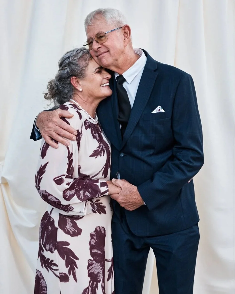 An elderly couple embracing, smiling, and sharing a tender moment, with a plain cream-colored backdrop.