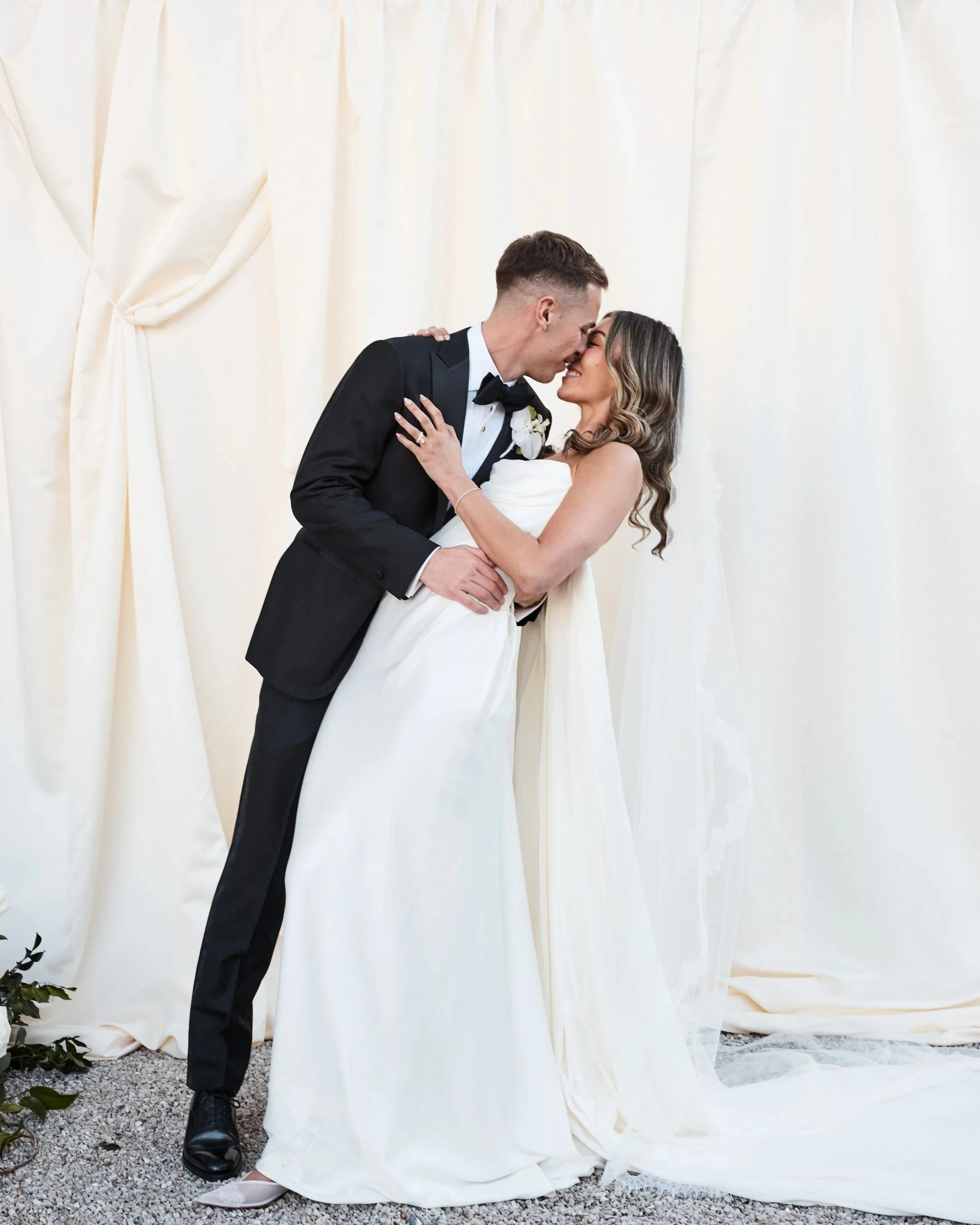 A bride and groom share a kiss during their wedding ceremony, with a cream-colored curtain backdrop.
