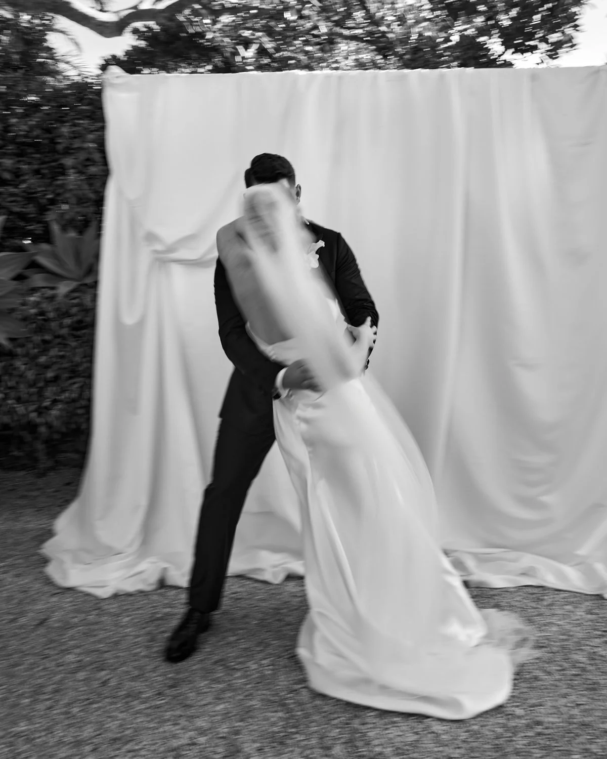 A bride and groom dancing outdoors in front of a white curtain backdrop, with trees in the background. The image is black and white.
