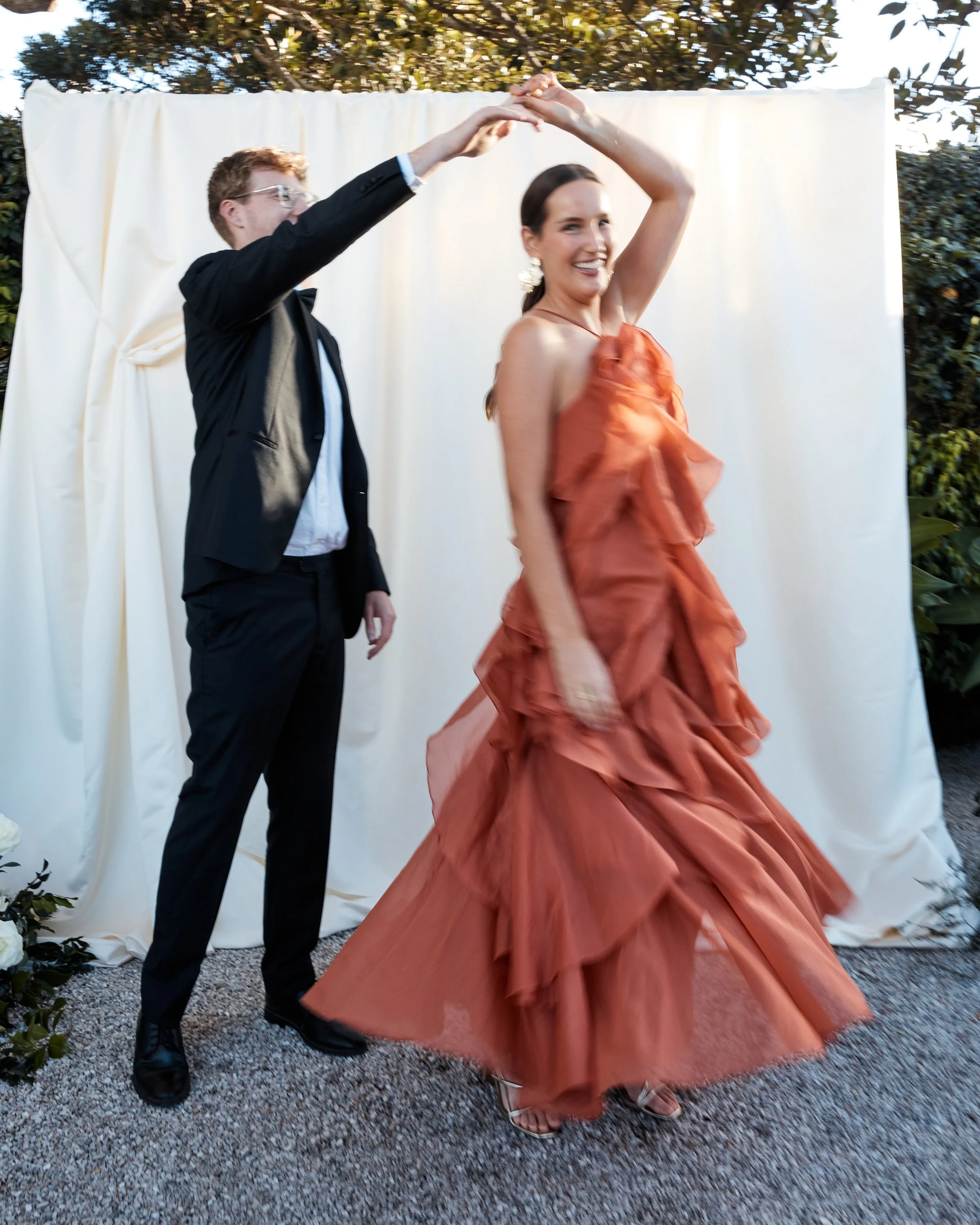 A man and woman dancing outdoors in front of a white backdrop, with the woman wearing a ruffled orange gown and the man in a black suit and glasses.