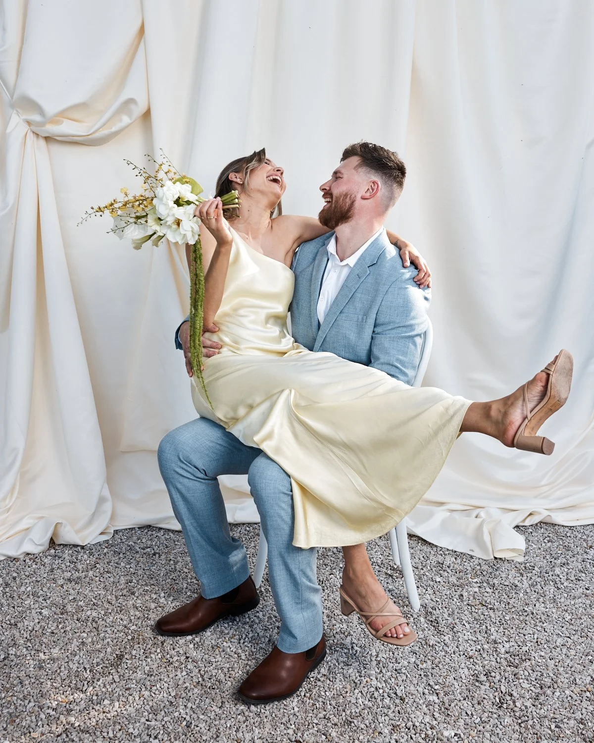 A couple celebrating their wedding, with the man sitting on a chair and carrying the woman on his lap, both smiling and looking at each other, in front of a white fabric backdrop. The woman is holding a bouquet of white flowers.
