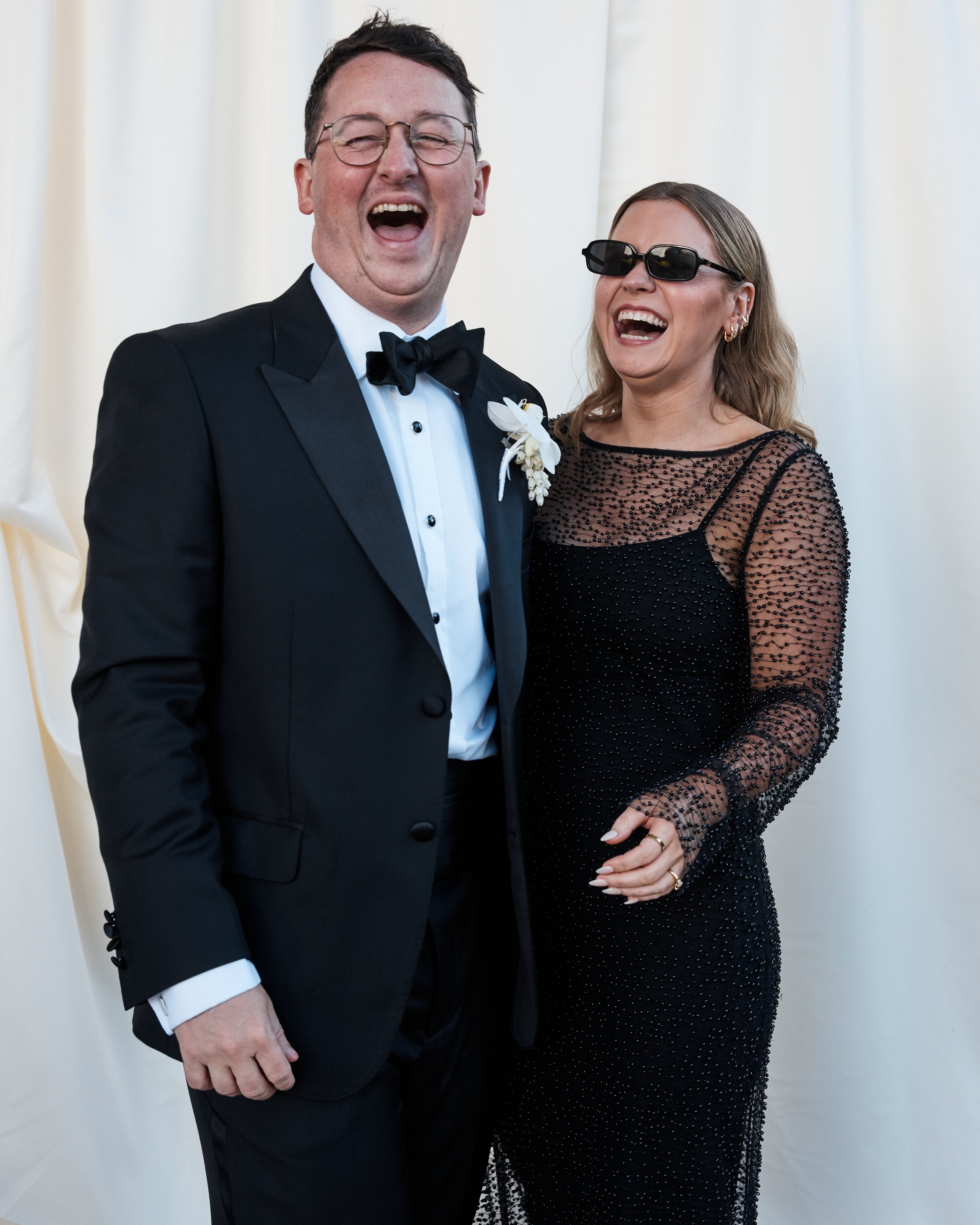 A man and woman laughing and smiling at a wedding, both dressed in formal attire with sunglasses.