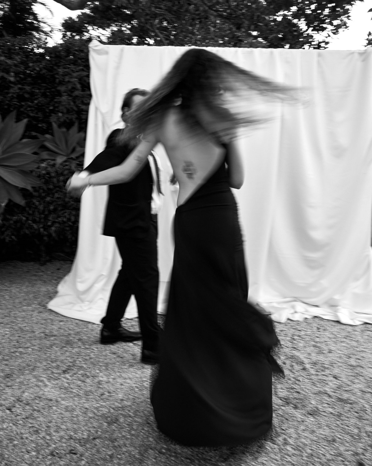 Black and white photo of two people dancing outdoors in front of a white curtain with trees in background. One person is wearing a dark suit and the other a long dark dress with flowing hair.