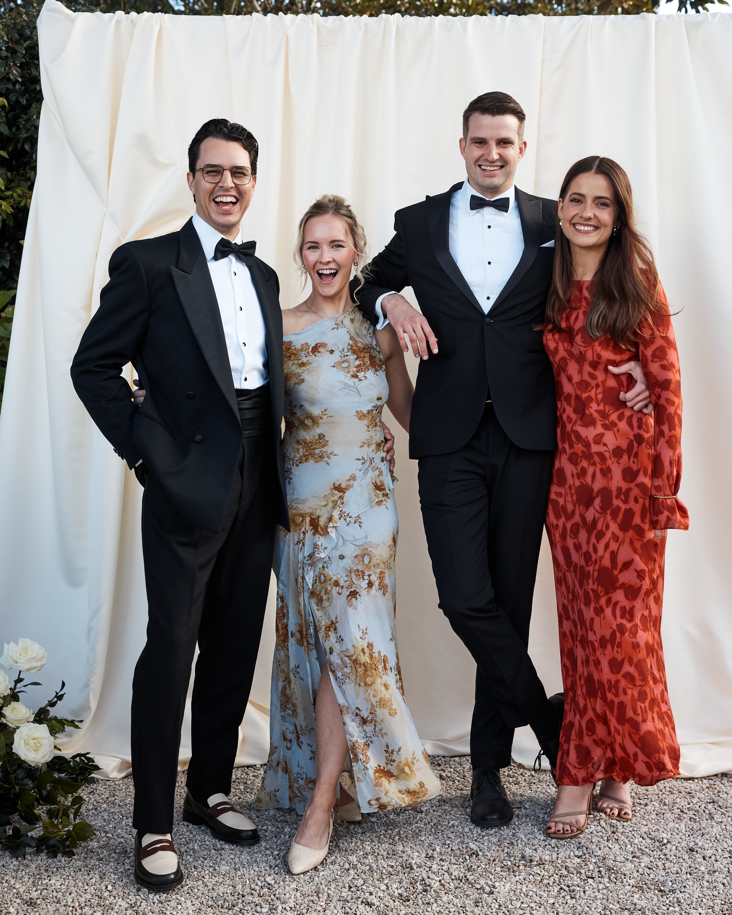 Four people standing together and smiling at an outdoor event, with a cream-colored fabric backdrop behind them. Two men in tuxedos and two women in elegant dresses.