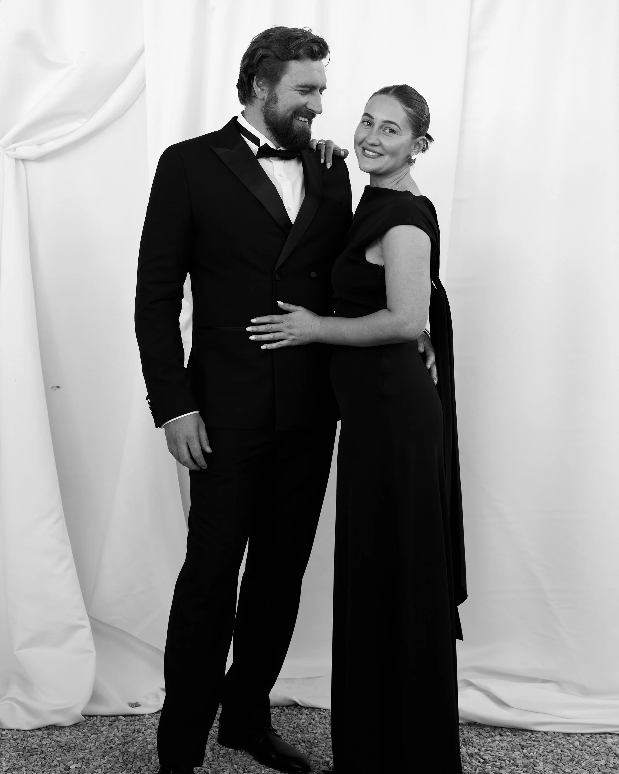 Black and white photo of a man and woman dressed in formal attire, smiling and standing close together in front of a white backdrop.