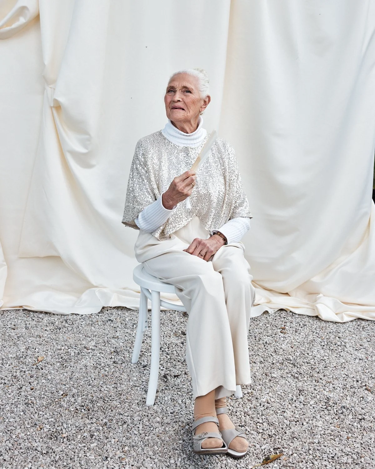 An elderly woman with white hair, dressed in a shiny silver top and white pants, sitting on a white stool against a plain white draped background, holding a small object in her right hand.