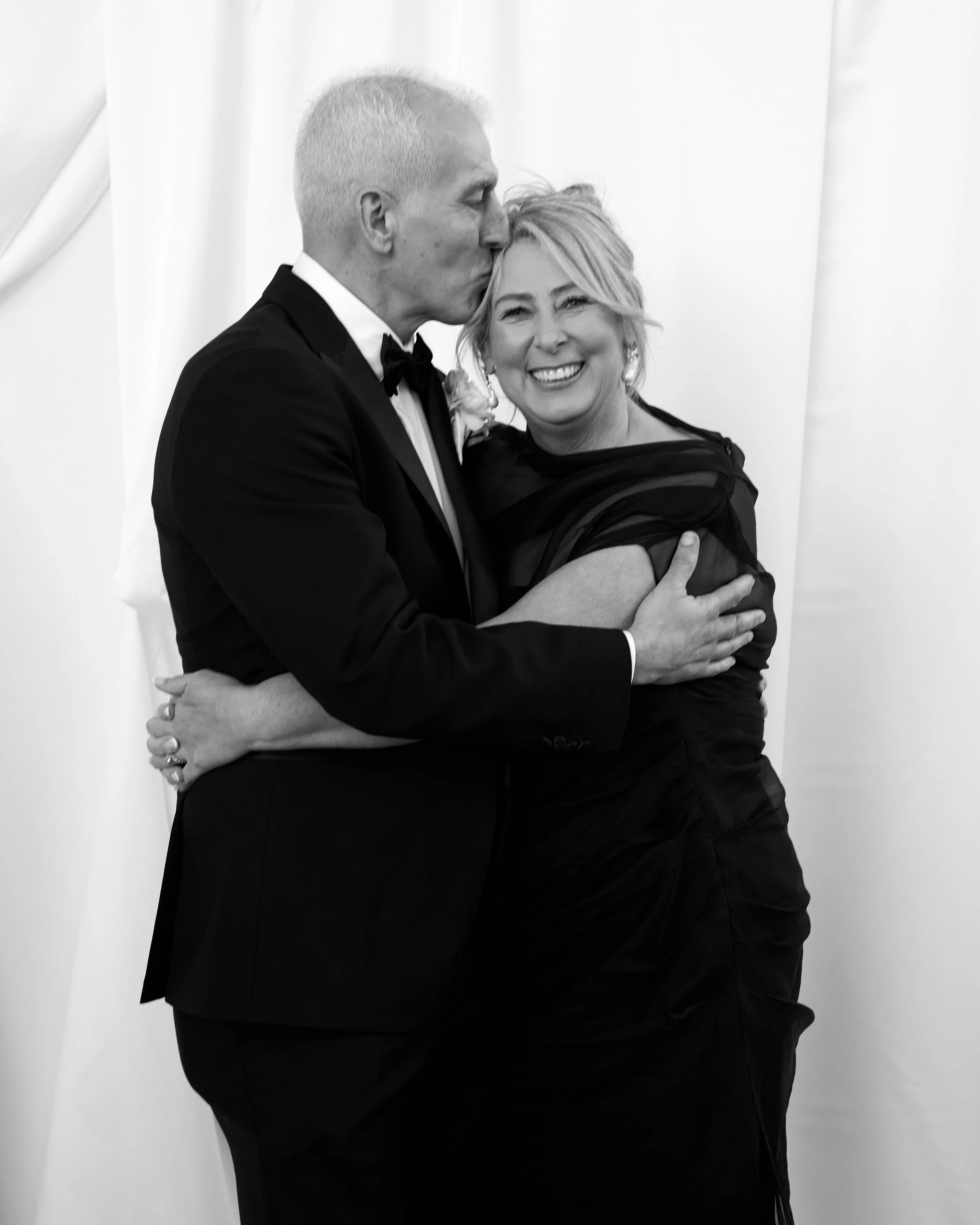 A man in a tuxedo kisses a woman in a black dress on the head at a formal event, both smiling.