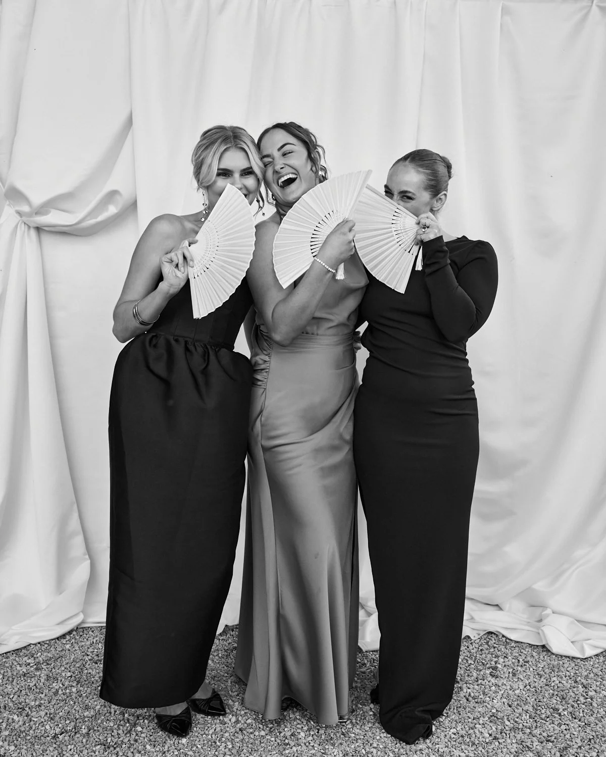 Three women in formal dresses holding fans, smiling and laughing, standing in front of a light-colored curtain.