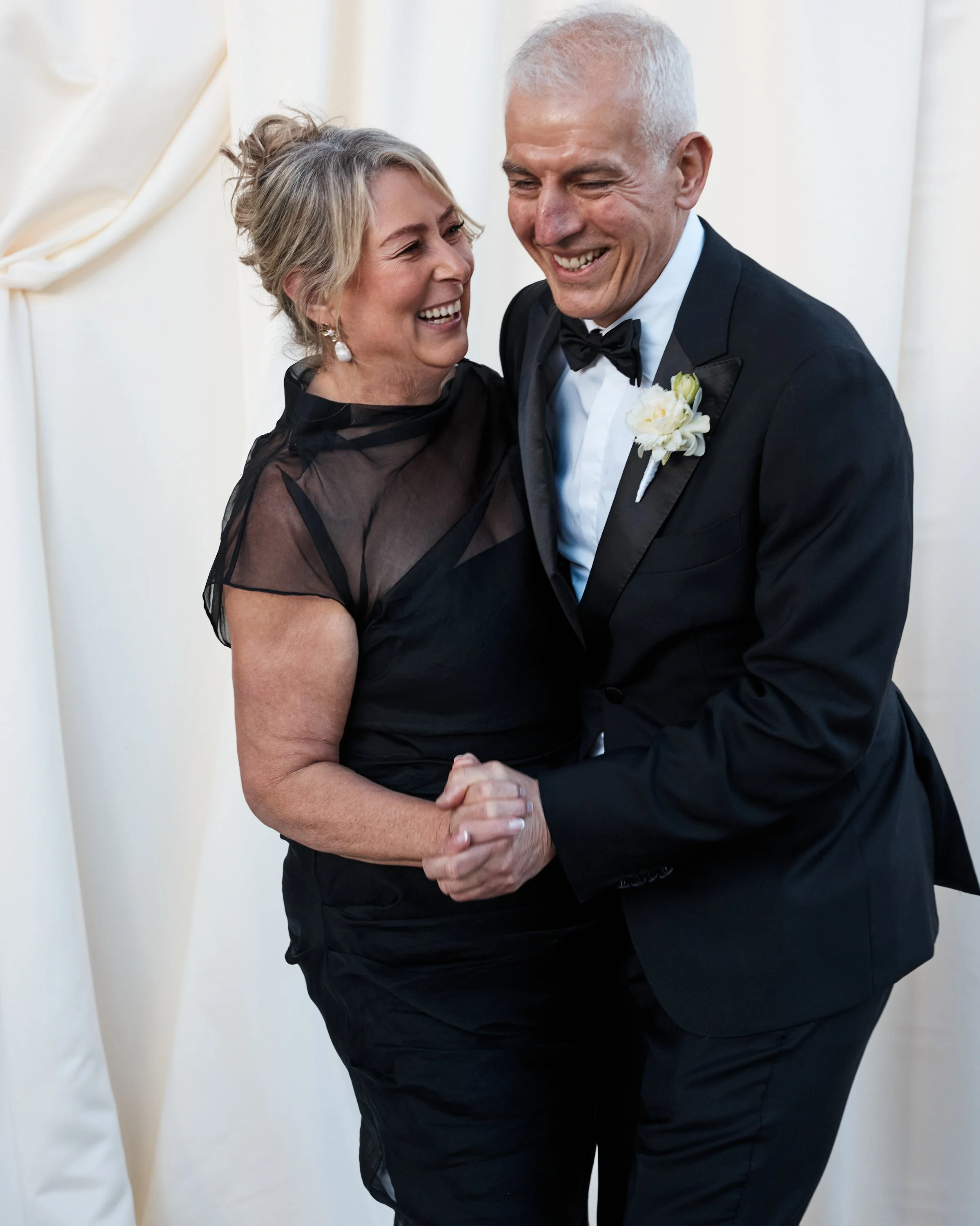 A smiling woman and man dressed in formal attire dance together, holding hands, at a special event or celebration.