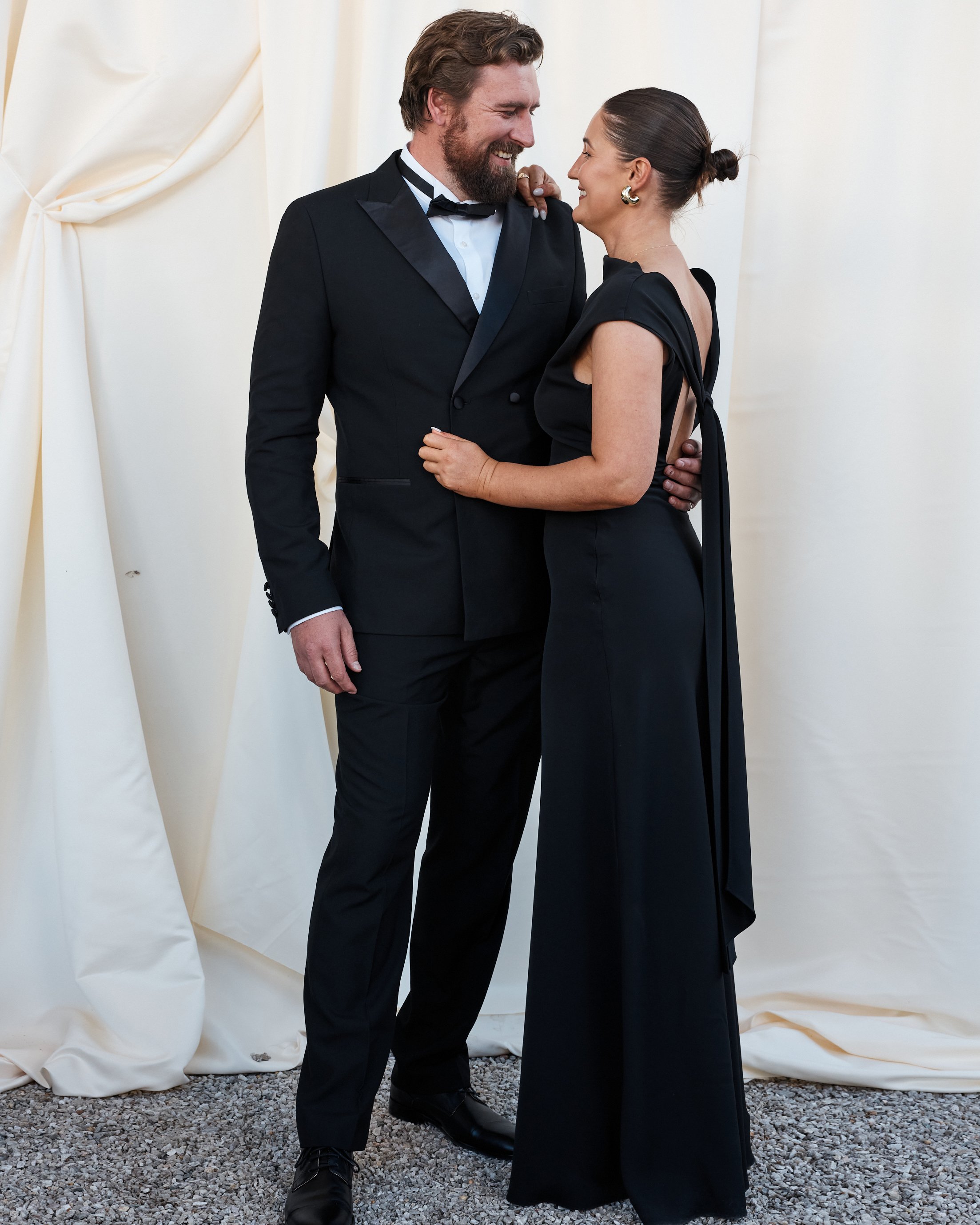 A couple dressed in formal black attire, smiling and looking at each other, standing close together against a cream-colored backdrop.