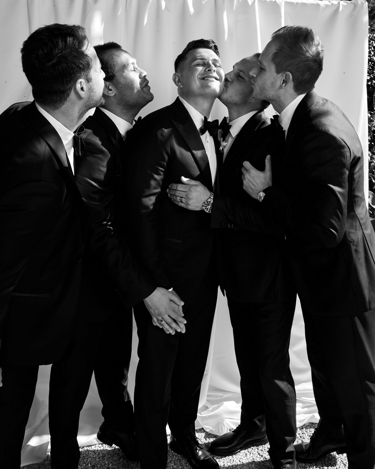 Six men in tuxedos celebrating at a wedding, with one man in the center smiling as he receives kisses from the others.
