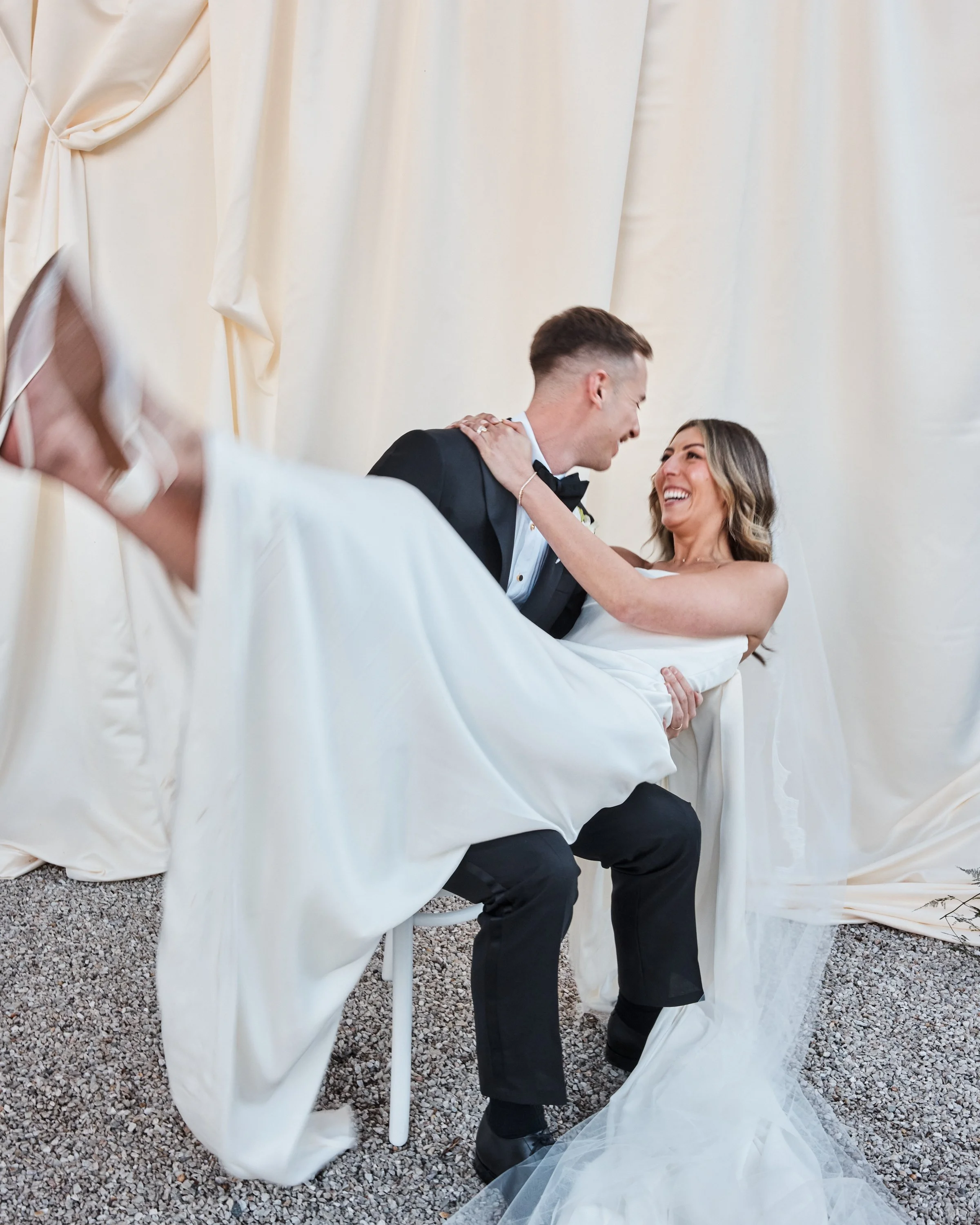 A bride and groom smiling and looking at each other, with the groom lifting the bride in his arms against a cream-colored curtain backdrop.
