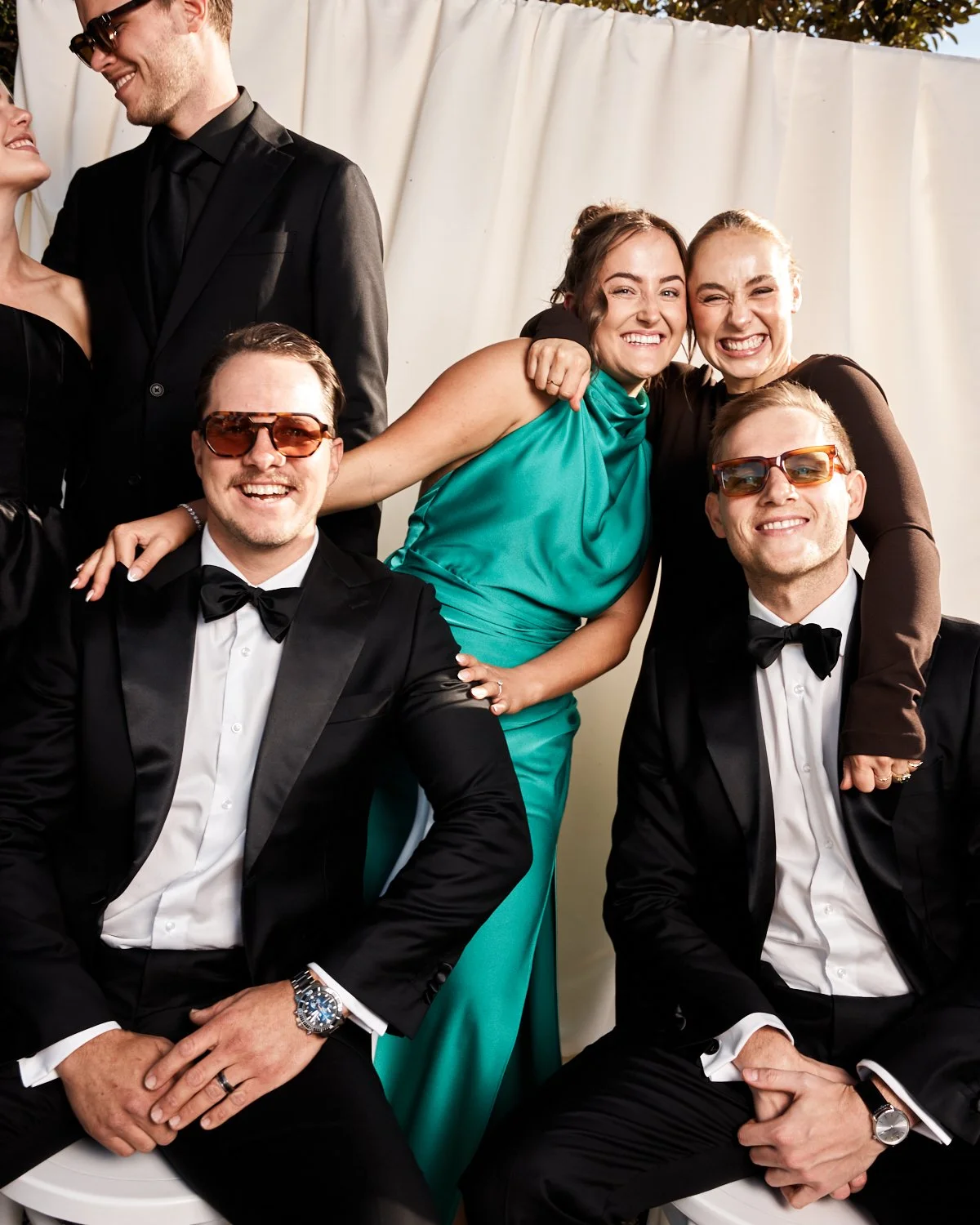Group of six people dressed in formal attire, smiling and posing together at an event, with a backdrop of a white curtain and outdoor trees.