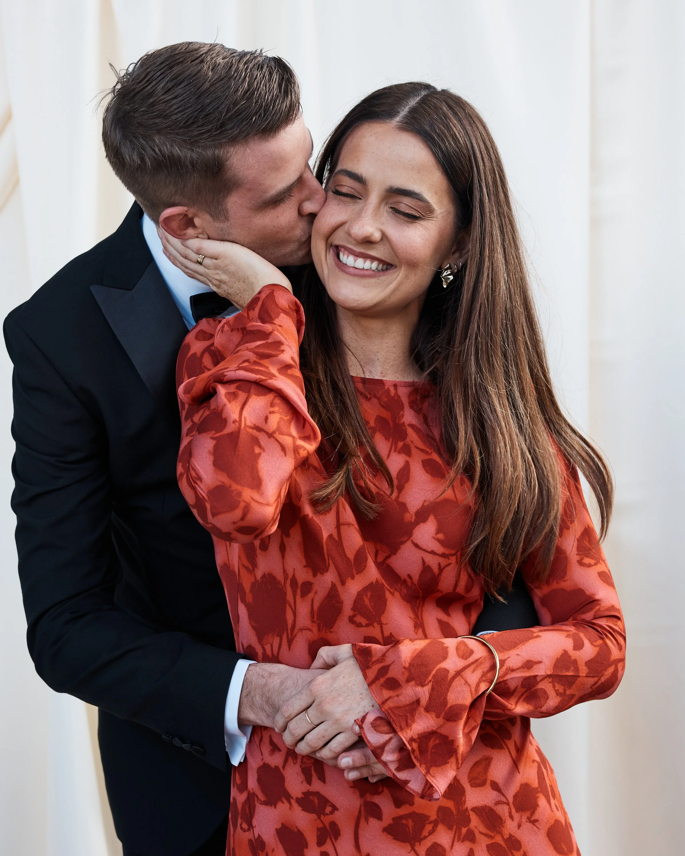 A man in a black tuxedo kisses a woman in a red floral dress on the cheek, she smiles with eyes closed.