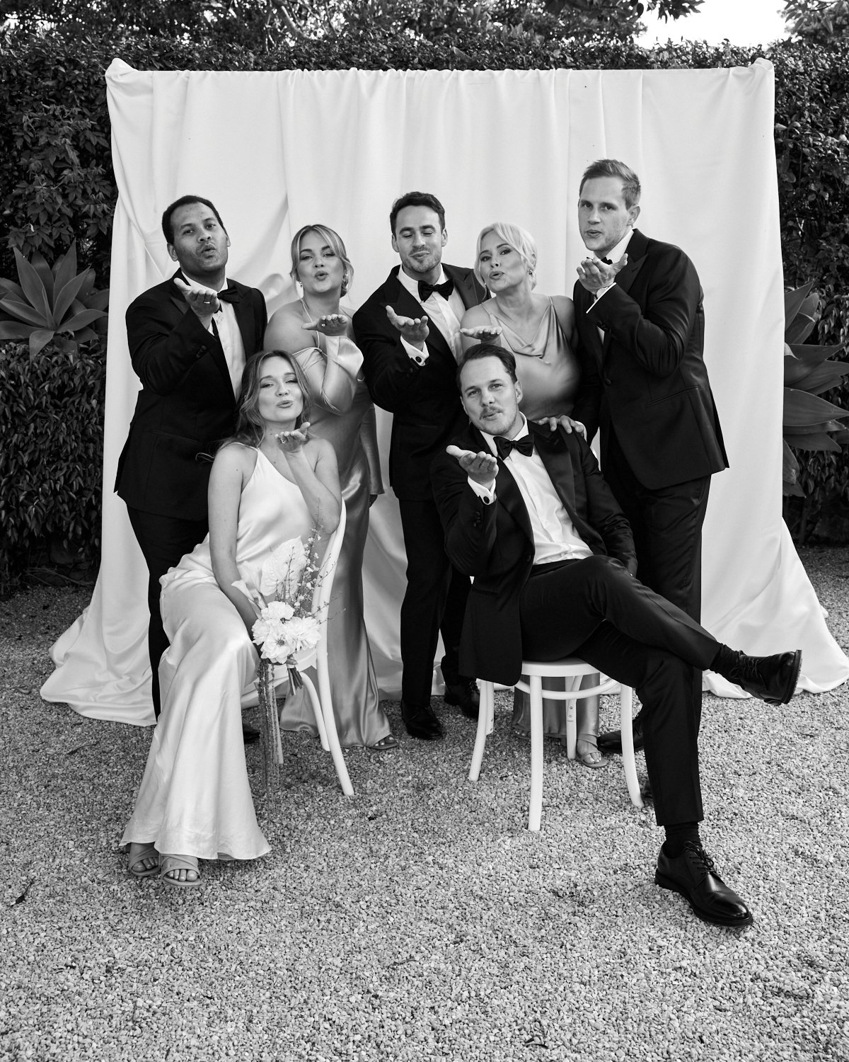 Group of seven people, dressed in formal attire, posing outdoors in front of a white curtain, with some blowing kisses.