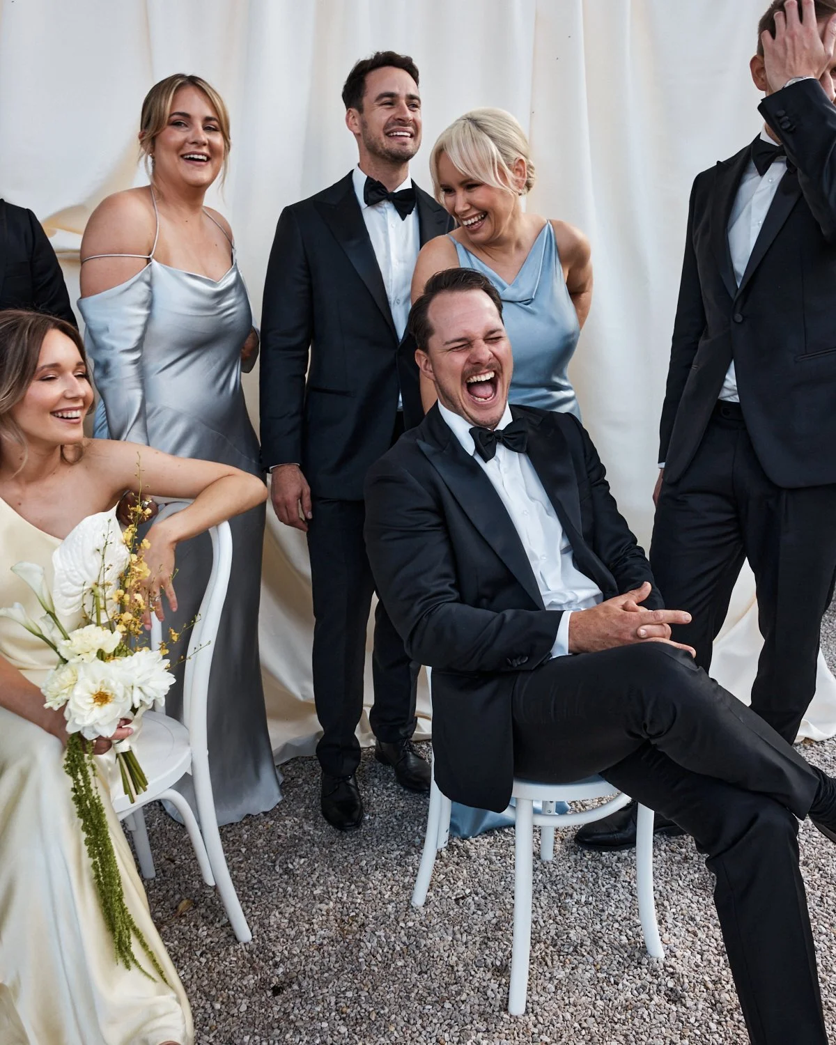 A group of people dressed in formal attire, smiling and laughing at a celebratory event, with some standing and one man seated, holding a small object, in front of a white curtain.