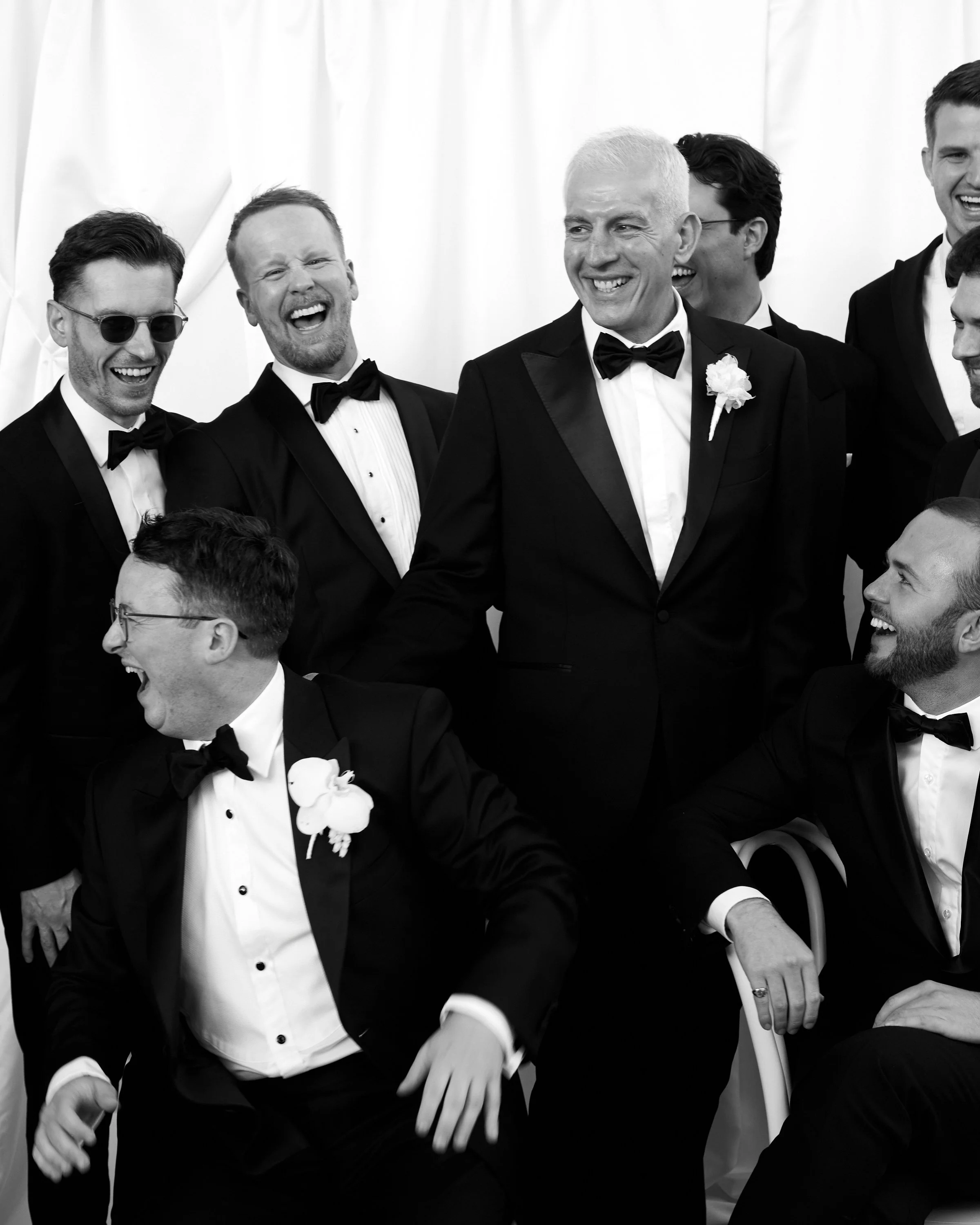 Group of men in formal tuxedos and bow ties, some with flowers, laughing and smiling at a celebration or wedding.
