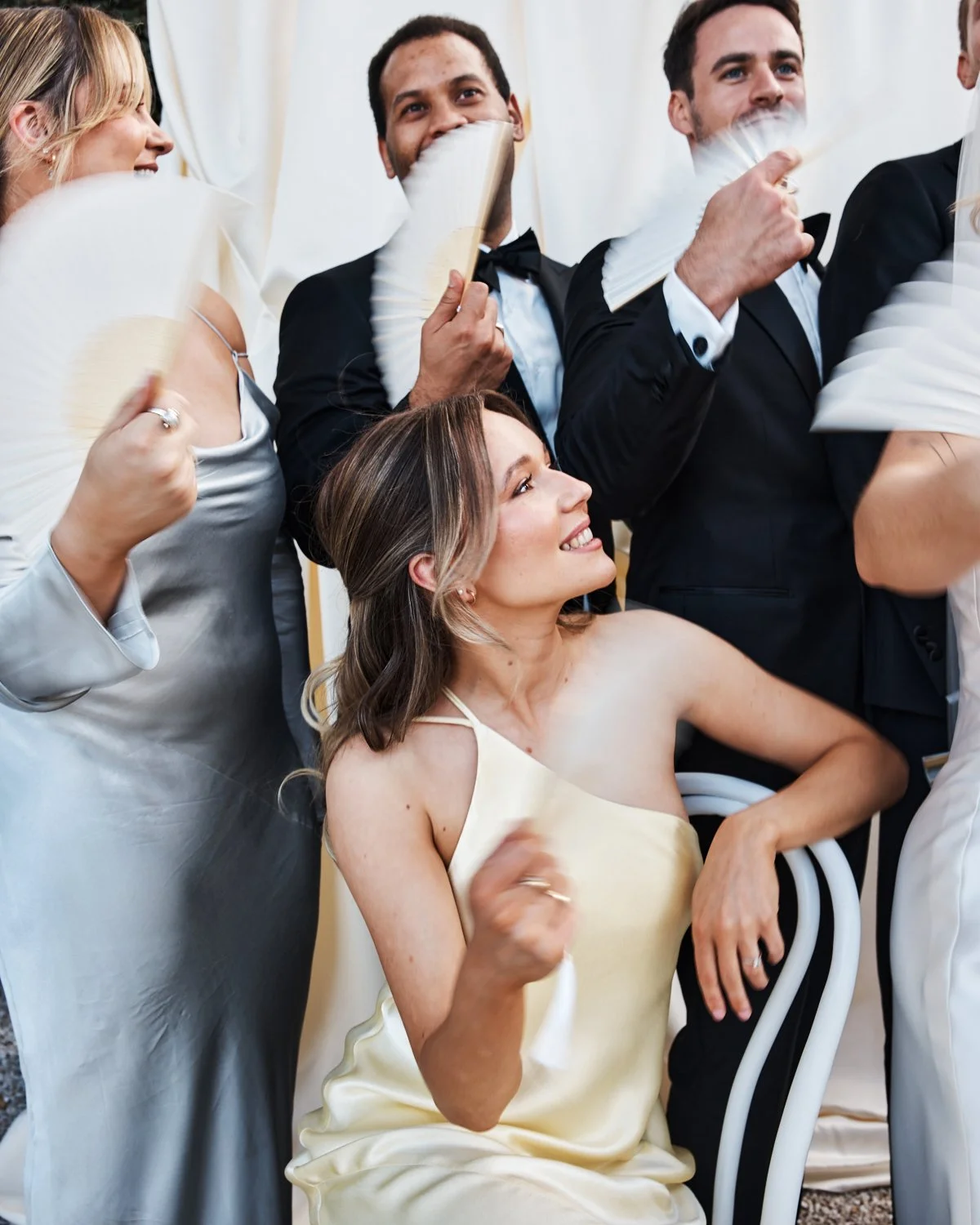 People at a social event, with some standing and one woman seated, smiling and holding a fan, dressed in formal or semi-formal attire.