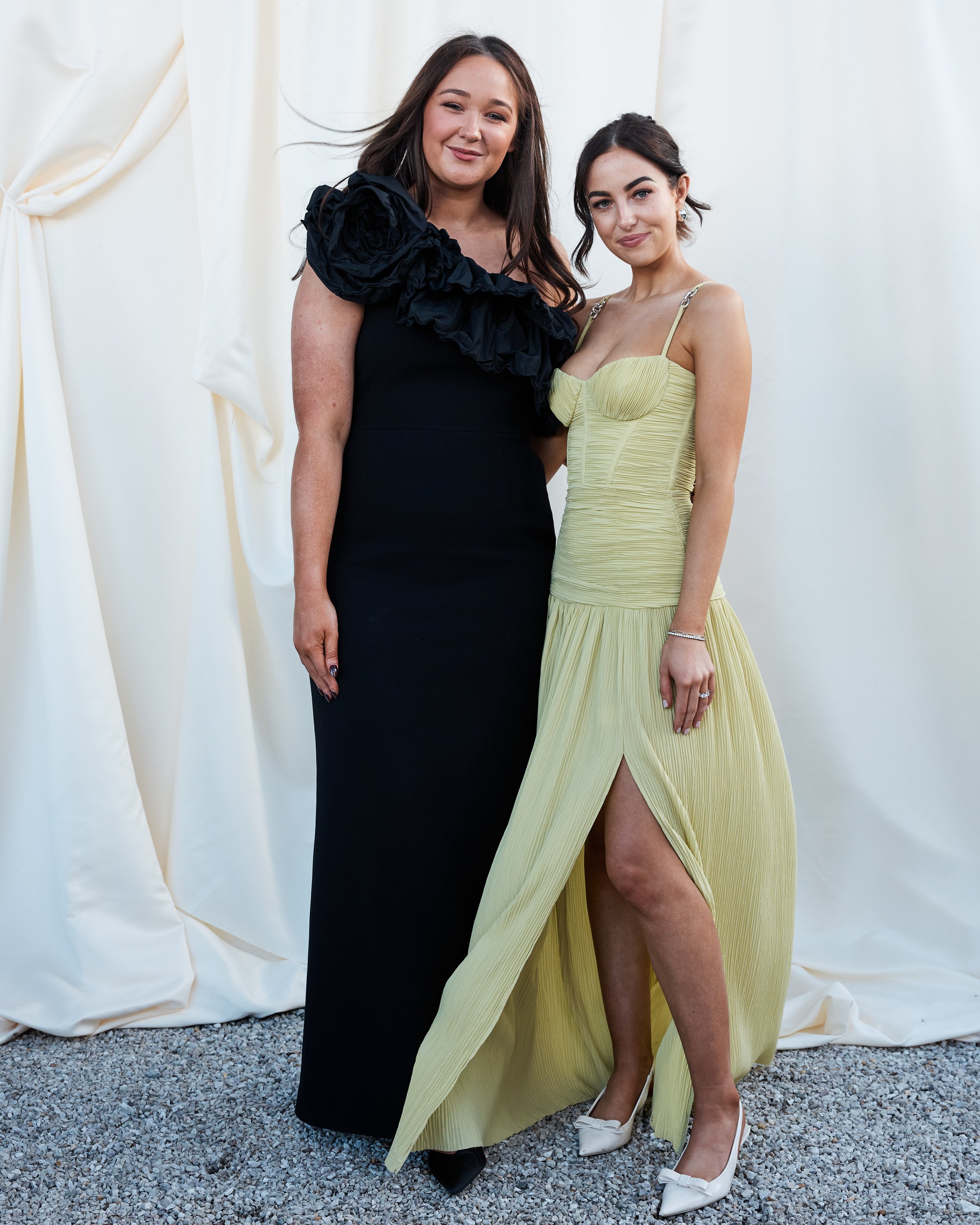 Two women standing together against a white backdrop, one in a long black dress with ruffled shoulder detail, the other in a long yellow dress with a high slit, both smiling at the camera.