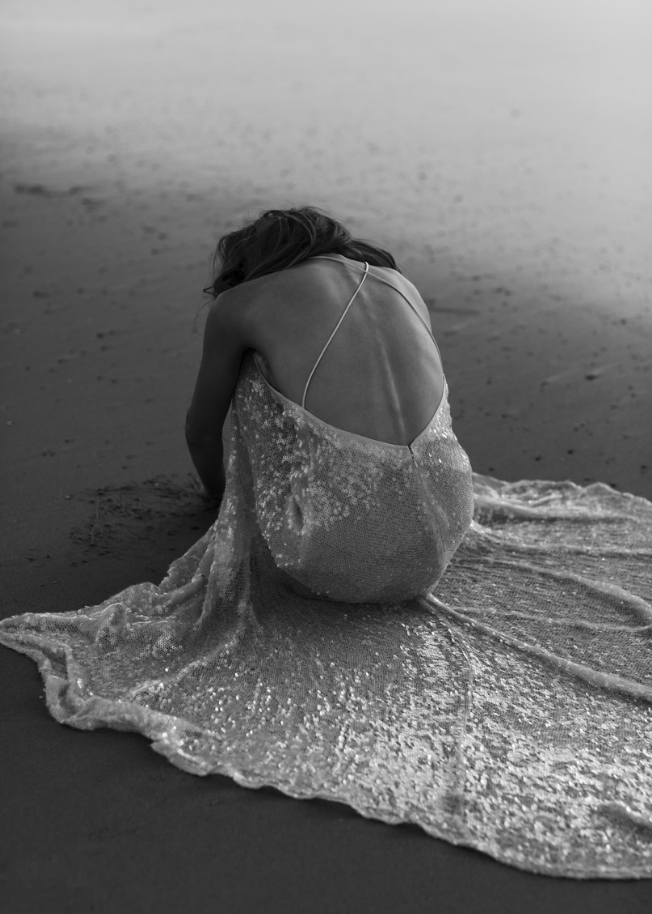 A woman sitting alone on a sandy beach in a sparkling dress, facing away from the camera, with her head bowed.
