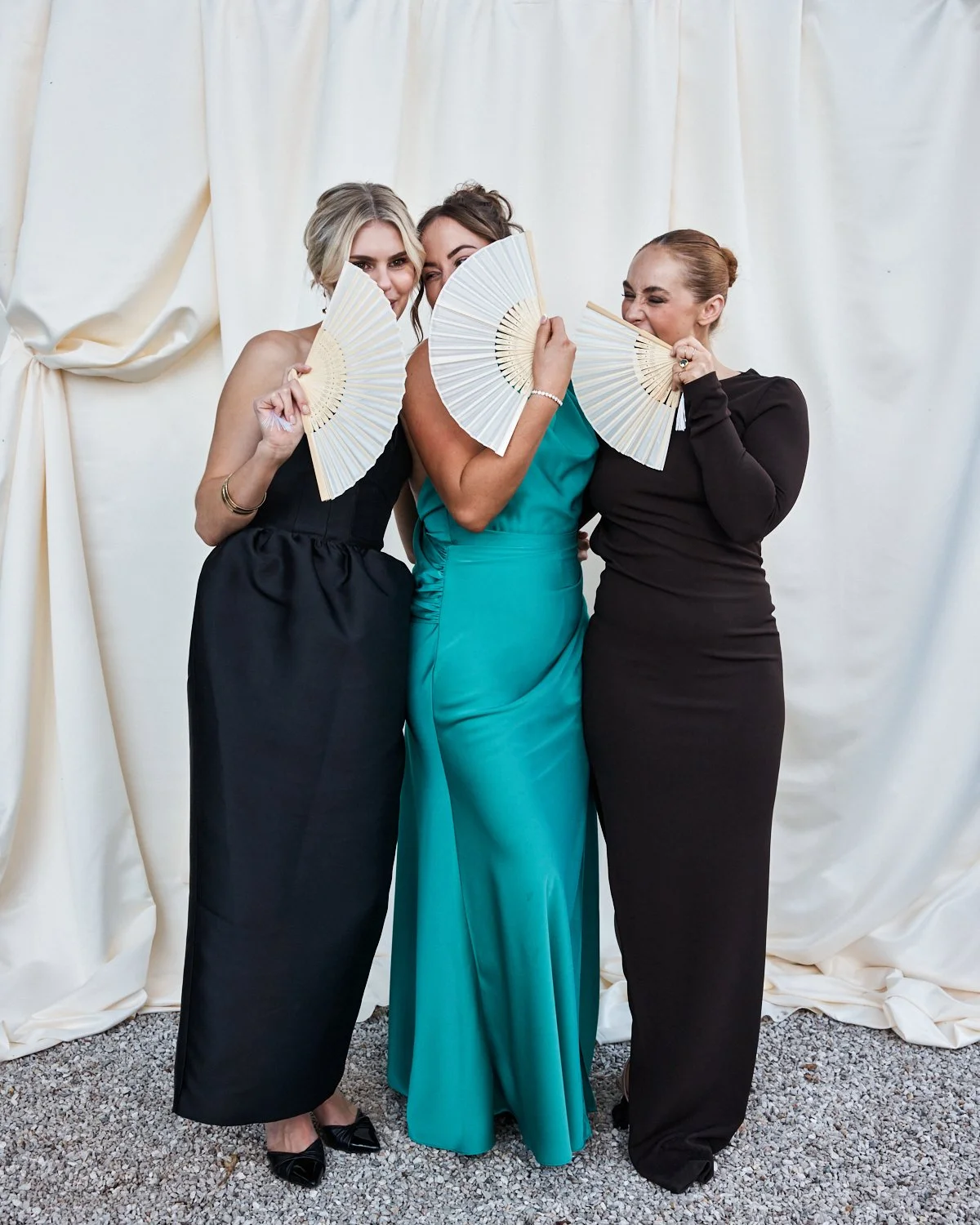 Three women in elegant dresses holding handheld fans, standing in front of a cream-colored curtain backdrop, smiling and posing playfully.