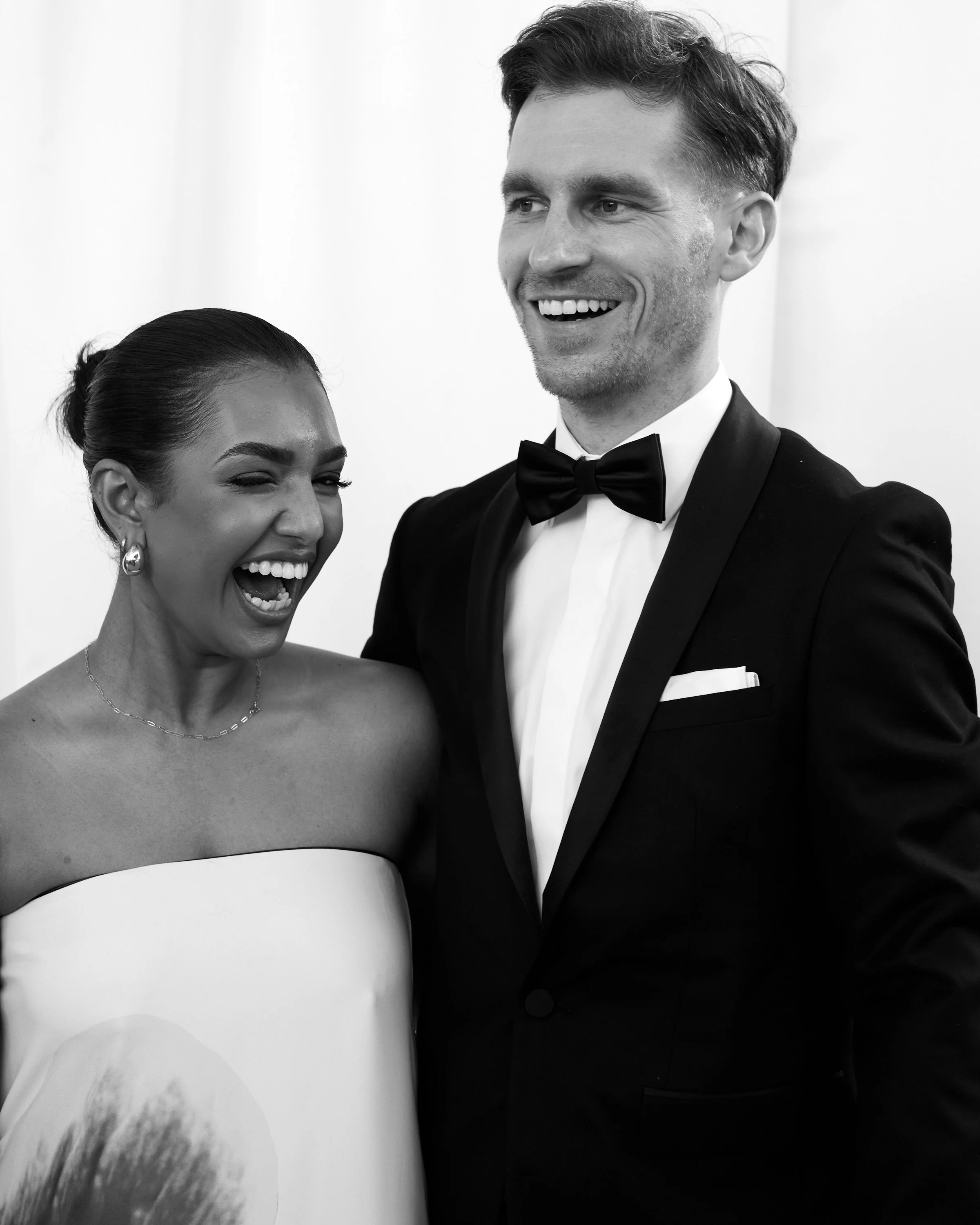 Man and woman in formal attire laughing and smiling, with the man wearing a tuxedo and bow tie, and the woman in a strapless dress with earrings.
